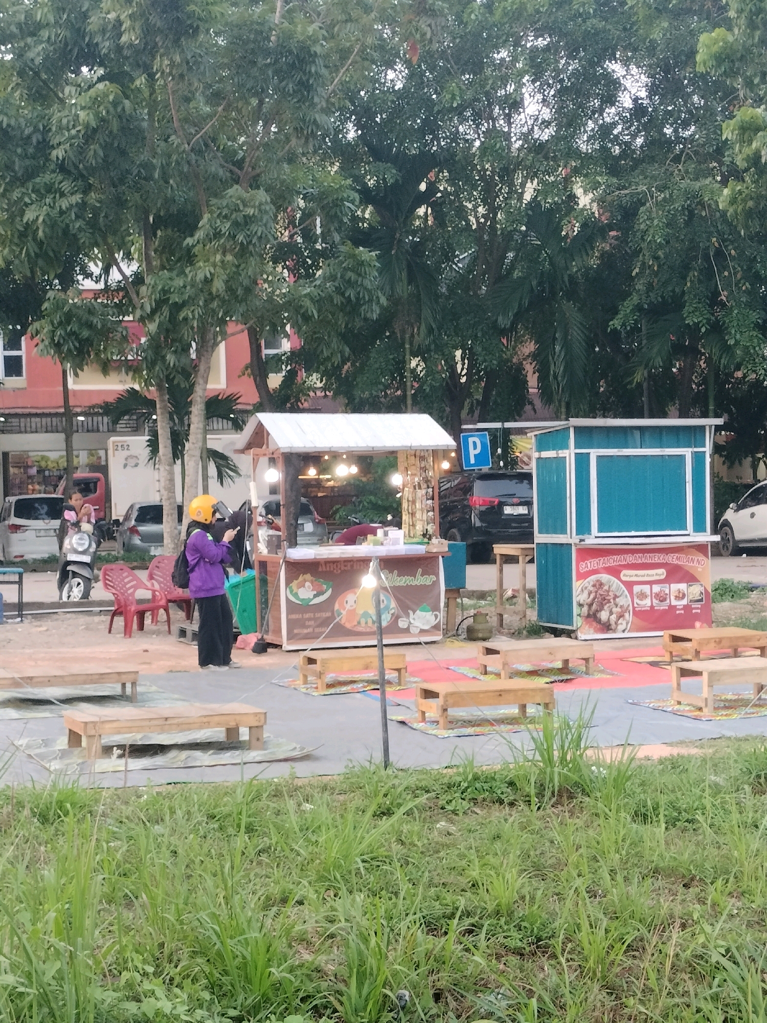 This image captures a street food stall located in the Batam City, Indonesia. The stall is set up outdoors, with a white canopy over the main counter. In front of the stall, several low wooden tables and benches are arranged on mats, suggesting a casual dining area.
A person wearing a purple jacket and a yellow helmet is standing near the stall, appearing to interact with the vendor or examine something in their hands. Another person is visible on a scooter in the background, to the left of the main stall.
Behind the stall, several vehicles are parked, including a black SUV with a blue parking sign above it. The background also shows buildings with red and beige facades, suggesting an urban or semi-urban environment. Lush green trees and foliage frame the scene, indicating that the area is outdoors and likely in a somewhat green part of the city. The lighting suggests it might be late afternoon or early evening. The overall scene depicts a typical street-side food vendor operation in Batam.