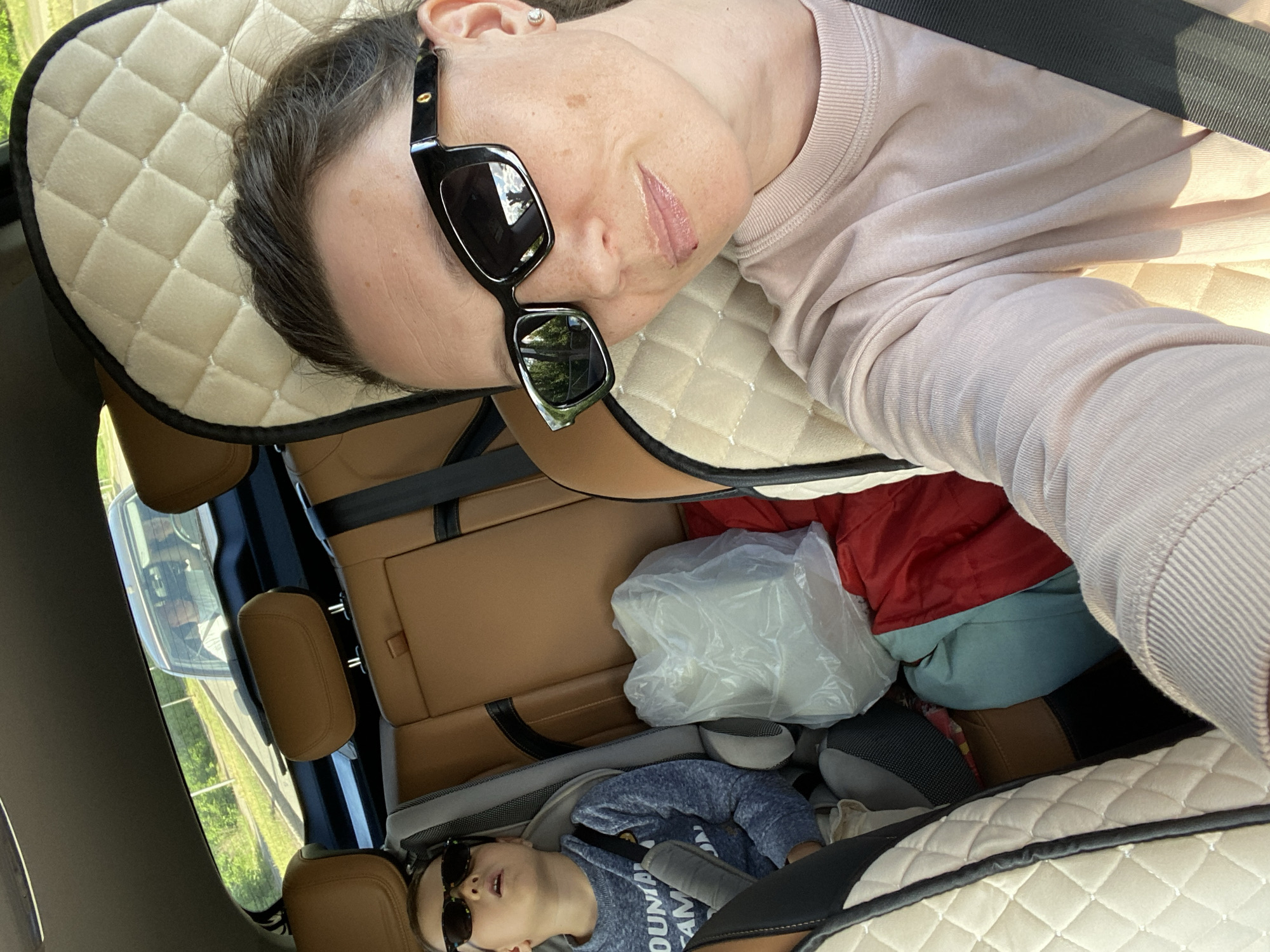 A woman with sunglasses lies in a car, resting her head on a beige, quilted headrest with black trim. She is wearing a light pink long-sleeved shirt. A child with sunglasses lies in the back seat of the car, secured by a seatbelt. Behind the child and between the front seats, there are items in the car, including a white plastic bag, a red jacket, and other clothing. The car's interior is tan and black. The exterior of the car is visible through the windows, revealing trees and a street. Based on the window reflection, the location is Chaikovsky, Russia.