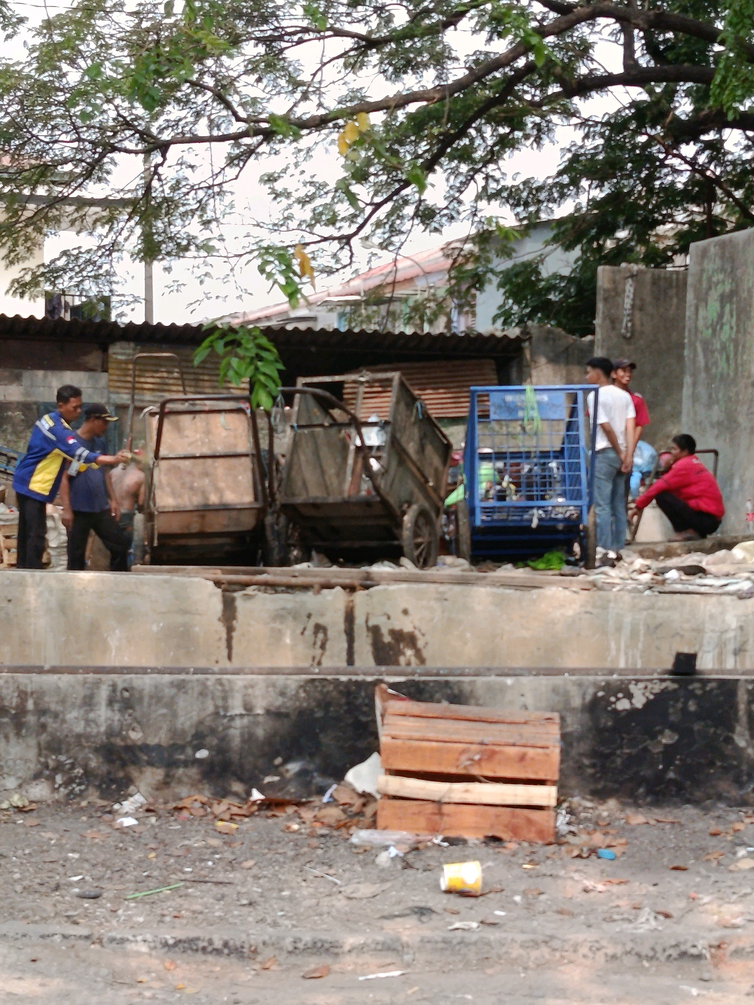 The image depicts a scene in what is identified as the city of Bekasi, Indonesia. In the foreground, a concrete barrier runs horizontally across the frame, with a littered ground surface beyond it. The ground is covered with debris, including leaves, paper, and plastic. A wooden crate is stacked on the ground.
In the middle ground, several individuals are present. On the left, two men stand near a large, wooden cart. One man, wearing a blue and yellow jacket, appears to be pointing or gesturing. The second man, wearing a dark shirt and cap, stands beside him. Further to the right, a blue metal cart is visible, containing some items. Several other people are standing or squatting in the background, some near a wall. The environment appears to be somewhat urban and industrial, with corrugated metal structures visible behind the carts. Lush green trees with dark branches extend from the top of the frame, suggesting an outdoor setting.
