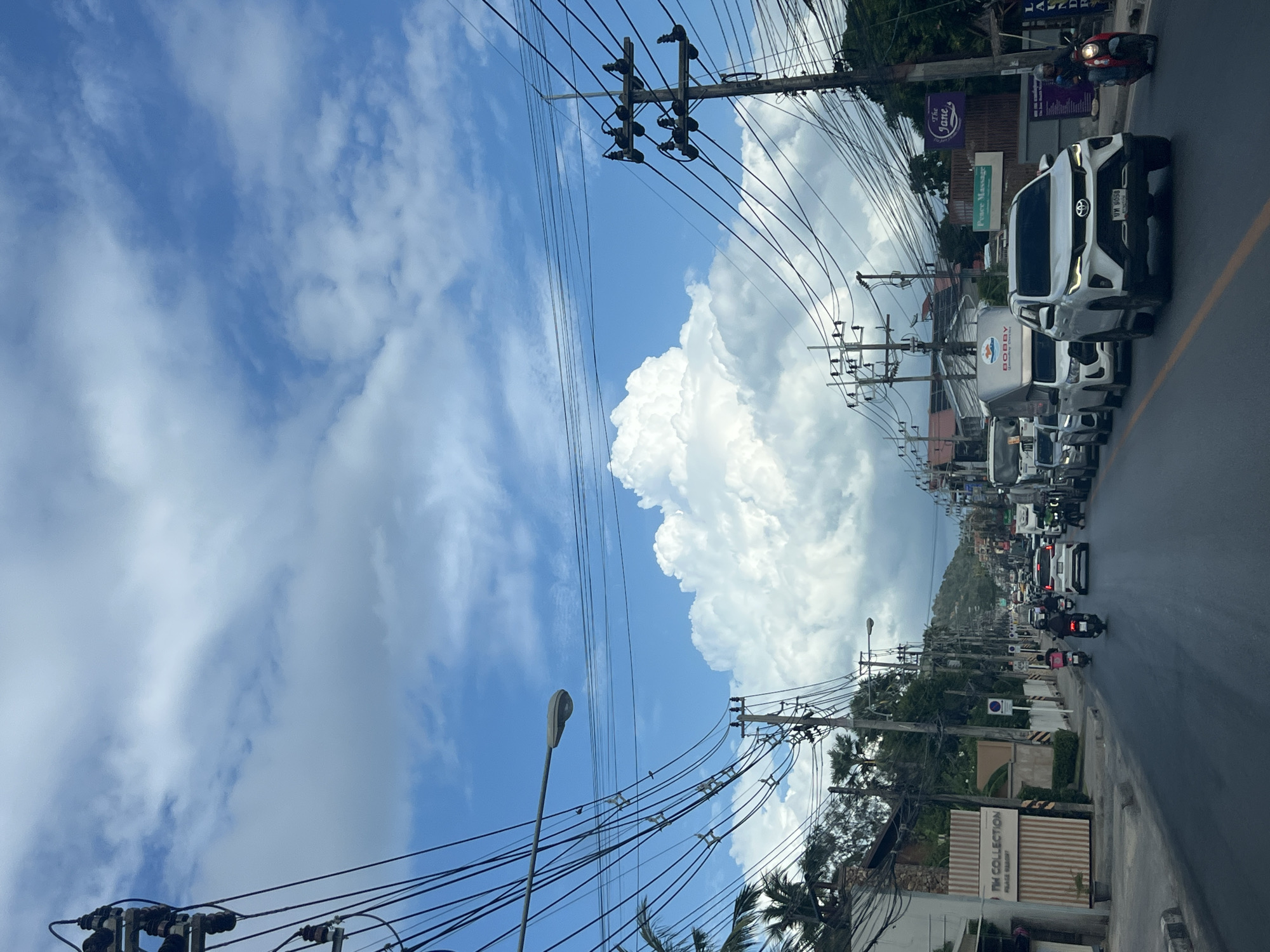 The image depicts a street scene in a city with a blue sky filled with large, fluffy white clouds. The road is lined with buildings and vegetation on both sides, with numerous power lines and utility poles dominating the foreground and midground. Vehicles, including cars and motorcycles, are present on the road, suggesting moderate traffic. The architectural style of the buildings appears to be a mix of modern and perhaps older structures. The overall atmosphere is one of a typical urban environment on a cloudy day.