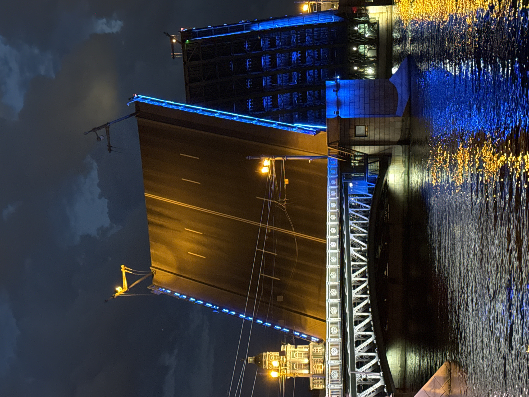 This image depicts a nighttime scene of a drawbridge in Saint Petersburg, Russia, likely during the White Nights season due to the dramatic illumination and dark sky. The central focus is the Palace Bridge (Dvortsovy Most) with its spans raised, allowing passage for ships. One span of the bridge is tilted upwards at a steep angle, revealing its understructure and the roadway. This span is illuminated with warm yellow lights, highlighting the white dividing lines on the asphalt. The other span is also raised and is adorned with vibrant blue lights that outline its structural elements, creating a striking contrast with the dark sky.
To the left, a portion of a historic building, possibly part of the Rostral Columns or a nearby administrative building on Vasilyevsky Island, is visible, illuminated by yellow streetlights. This building has a domed roof and ornate architectural details. In the foreground, the dark waters of the Neva River reflect the colorful lights from the bridge and the city, creating shimmering patterns on the surface. The sky is filled with dark, dramatic clouds, adding to the atmospheric mood of the scene. There are no people visible in the image, and the scene is static, capturing the moment the bridge is in its raised position. The overall impression is one of grandeur and nighttime city activity, with the illuminated bridge serving as a prominent landmark.