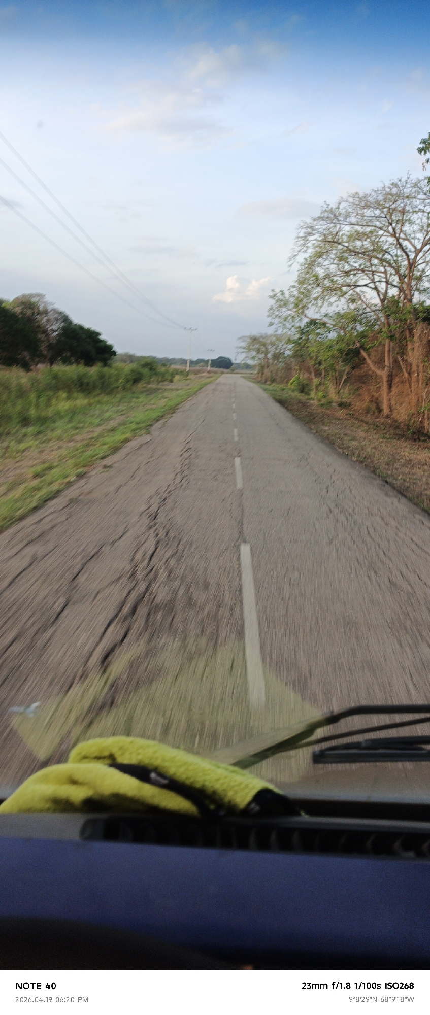 The image shows a view from the windshield of a vehicle traveling down a rural road. The road is cracked and has a faded white line marking the center. On either side of the road, there is vegetation and trees. Power lines run alongside the road. In the foreground, a yellow cloth rests on the dashboard of the vehicle. The photo information indicates it was taken with a 23mm lens at f/1.8, 1/100s, ISO268, and the location is 9°8'29"N 68°9'18"W, which corresponds to La Cruz, Venezuela. The time stamp suggests it was taken at 06:20 PM on April 19, 2026. The sky is partially cloudy.