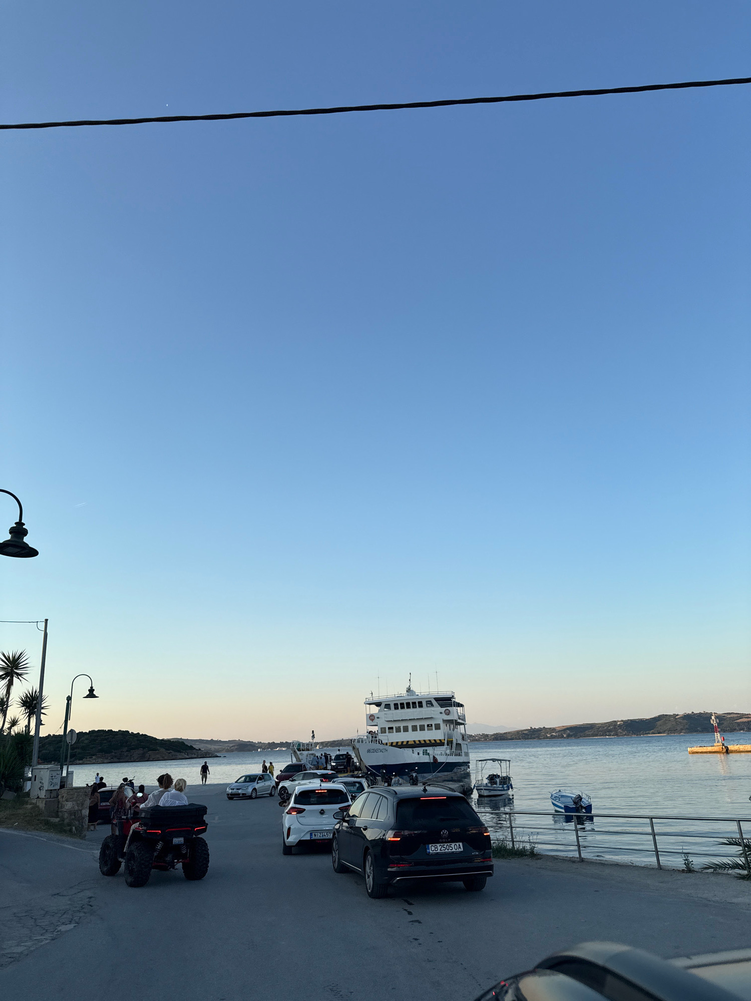 The image depicts a scene at a ferry terminal in Ammouliani, Greece, likely during the late afternoon or early evening, given the soft, warm light and clear sky. A large white ferry is docked, with its nameplate visible as "BEOCETAKETH". Several cars, including a black Volkswagen SUV with the license plate "CB 2505 OA", a white car with license plate "NI Z14 57", and a smaller white car are lined up on the asphalt. In the foreground, a red ATV with two passengers is moving away from the camera.
To the left, palm trees and a streetlamp are visible along a paved road. Several people are gathered near the water's edge, some walking and others standing. The sea is calm, reflecting the sky, and several small boats are moored or anchored. In the distance, a hilly, green coastline is silhouetted against the horizon. The overall impression is one of a relaxed, end-of-day atmosphere at a coastal transportation hub.