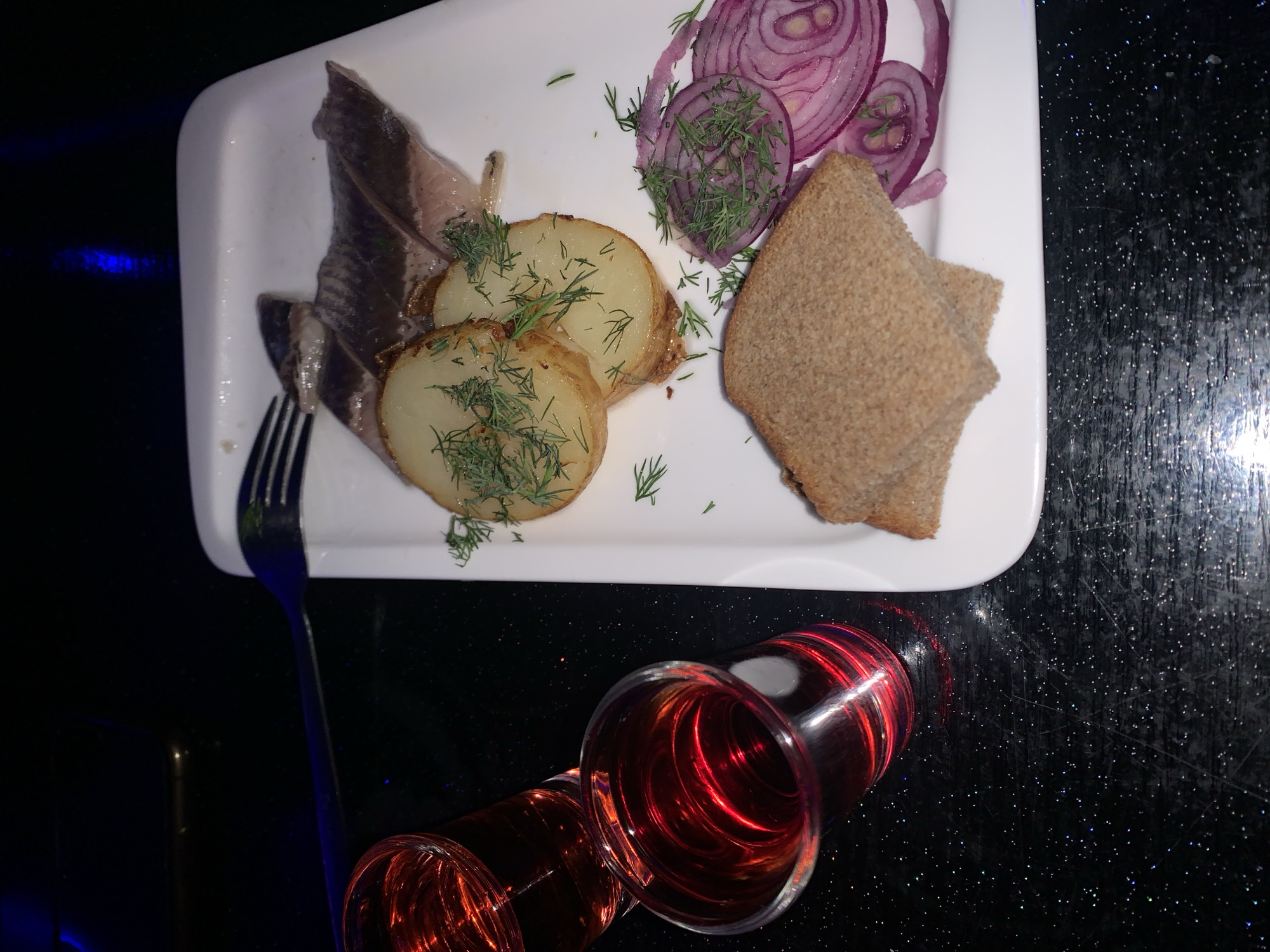 The image shows a plate of food with slices of herring, boiled potatoes, red onion rings, and rye bread, garnished with dill. Two shot glasses filled with a red liquid, likely vodka or liqueur, are placed to the left of the plate. The setting appears to be a bar or restaurant with a dark, reflective surface, possibly a table, that sparkles with small lights. The overall lighting is dim with blue and purple reflections, suggesting a nighttime ambiance. The food is arranged on a white rectangular plate. The scene is devoid of any people or visible interactions. The location is identified as Sinyavino, Russia.