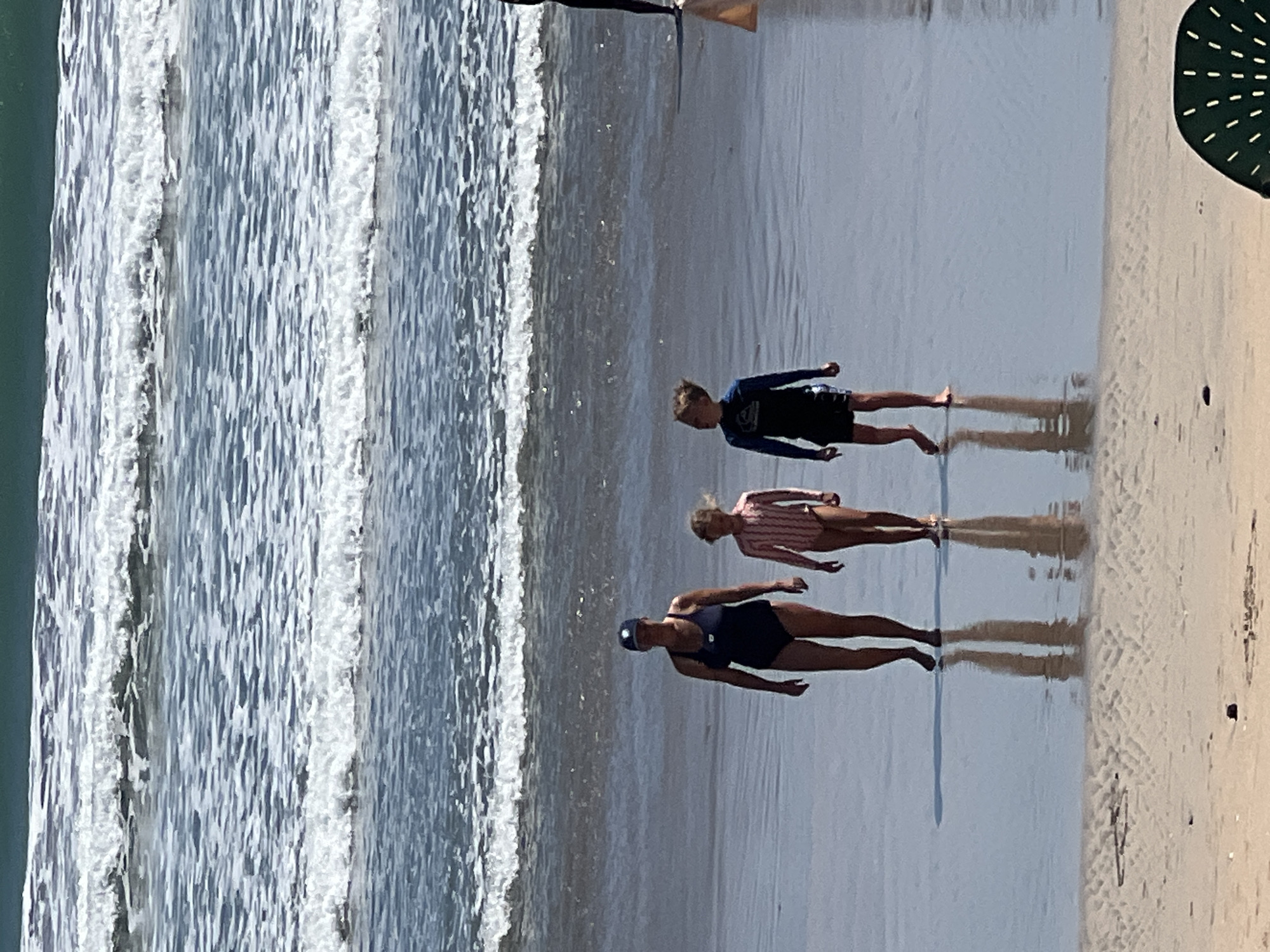 An overhead, full shot captures a family of three walking on a sandy beach. The family is positioned in the middle ground of the image, with the ocean in the background and a stretch of sand in the foreground. The ocean, with its waves, occupies the upper half of the image, displaying shades of blue and white. The sand is wet and reflects the sky and the figures.
On the left is a woman wearing a dark blue swimsuit and a navy blue baseball cap. To her right is a young girl in a pink and white striped swimsuit. To the girl's right is a young boy wearing a dark blue rash guard with a light blue logo and dark shorts. All three are walking away from the ocean towards the viewer. The boy is looking to his left. The woman appears to be looking forward. The girl's gaze is not discernible. Their reflections are visible in the wet sand. The foreground of the image shows dry sand with some pebbles. A dark green beach chair is partially visible in the bottom right corner. The scene is set in Kuta, Indonesia.
