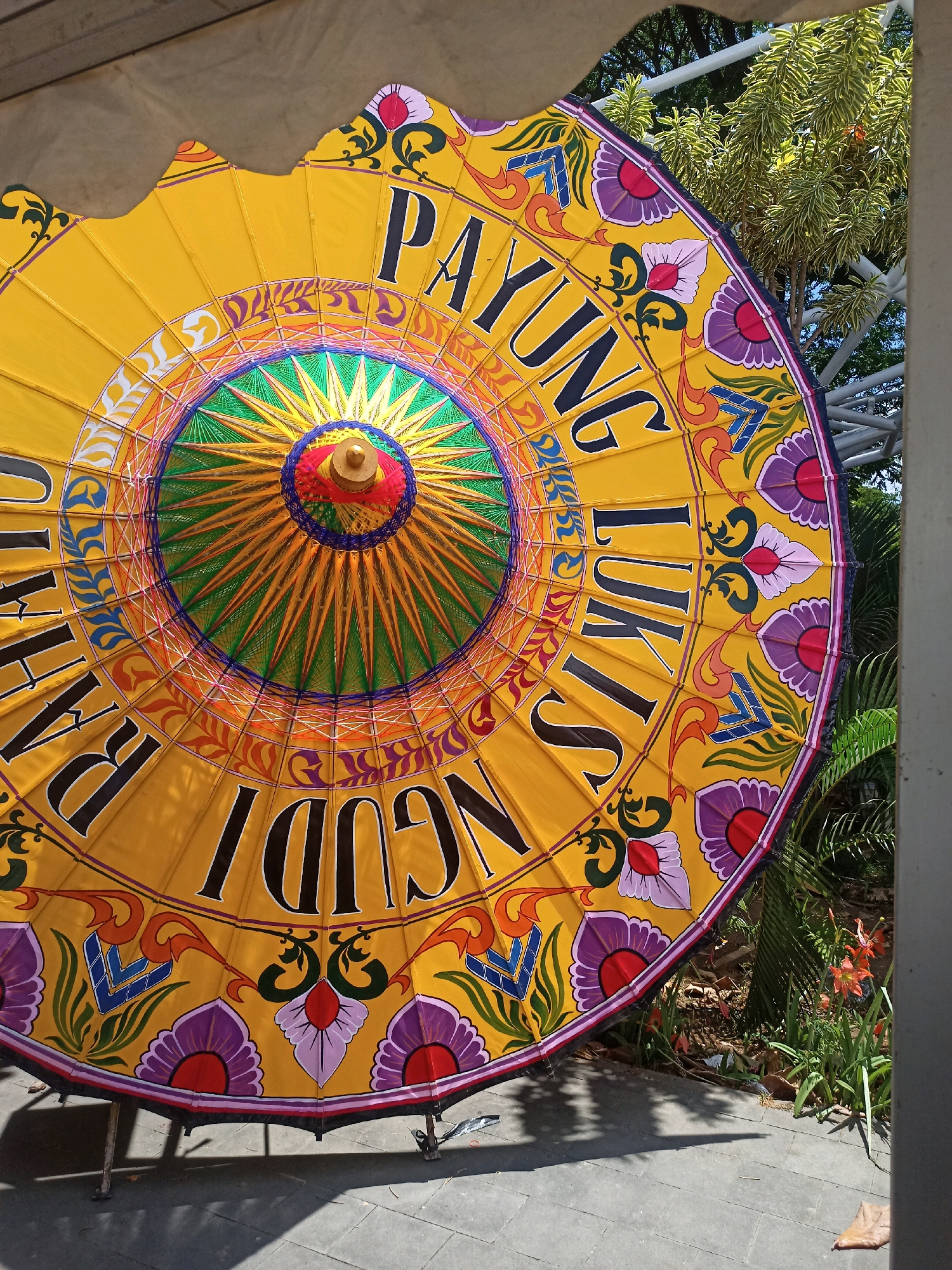 The image shows a close-up, partially angled view of a large, ornate, yellow umbrella, likely a decorative parasol. The umbrella is predominantly yellow with vibrant, colorful patterns and text. The text, written in black capital letters in a decorative font, appears to be in Indonesian. From what is visible, the text reads "PAYUNG" and "LUKIS" on the upper right portion, and "NGUDI" and possibly part of "SARI" on the lower left. The center of the umbrella features a radial design with green and orange spokes emanating from a red and blue circular hub, topped with a gold finial. The outer edge of the umbrella is decorated with stylized purple flowers with red centers, green leafy motifs, and orange and blue abstract designs. The umbrella is propped open and angled downwards, casting a shadow on the paved ground beneath it. The background includes lush green foliage, including palm-like leaves and some orange flowers, suggesting an outdoor setting. A metal structure, possibly part of a canopy or trellis, is visible in the upper right background. The paving stones appear to be square and light gray. This type of decorative umbrella, known as a "Payung Lukis" or painted umbrella, is a traditional craft associated with the city of Surakarta (also known as Solo), Indonesia. Surakarta is renowned for its rich cultural heritage, including its traditional crafts and artistic expressions, and these umbrellas are often displayed in cultural events, tourist areas, or as decorative elements within the city.