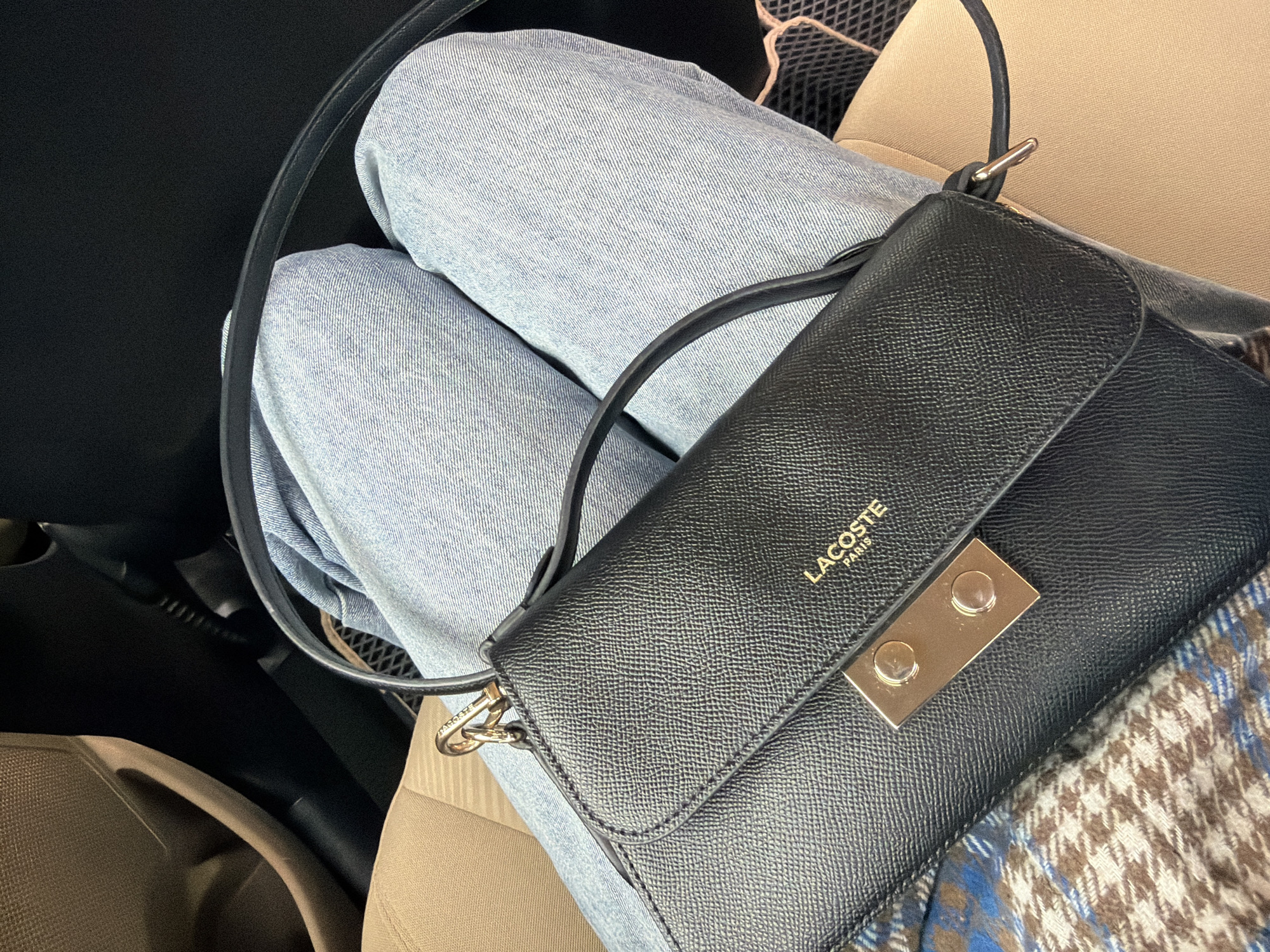 The image shows a person wearing light-wash jeans sitting in what appears to be a car. A black Lacoste handbag with a gold clasp and the Lacoste logo is positioned on their lap. Part of a patterned scarf or blanket is visible beneath the bag. The car interior includes beige upholstery and dark dashboard elements. The location in the city of Yerevan, Armenia, cannot be confirmed from the image.