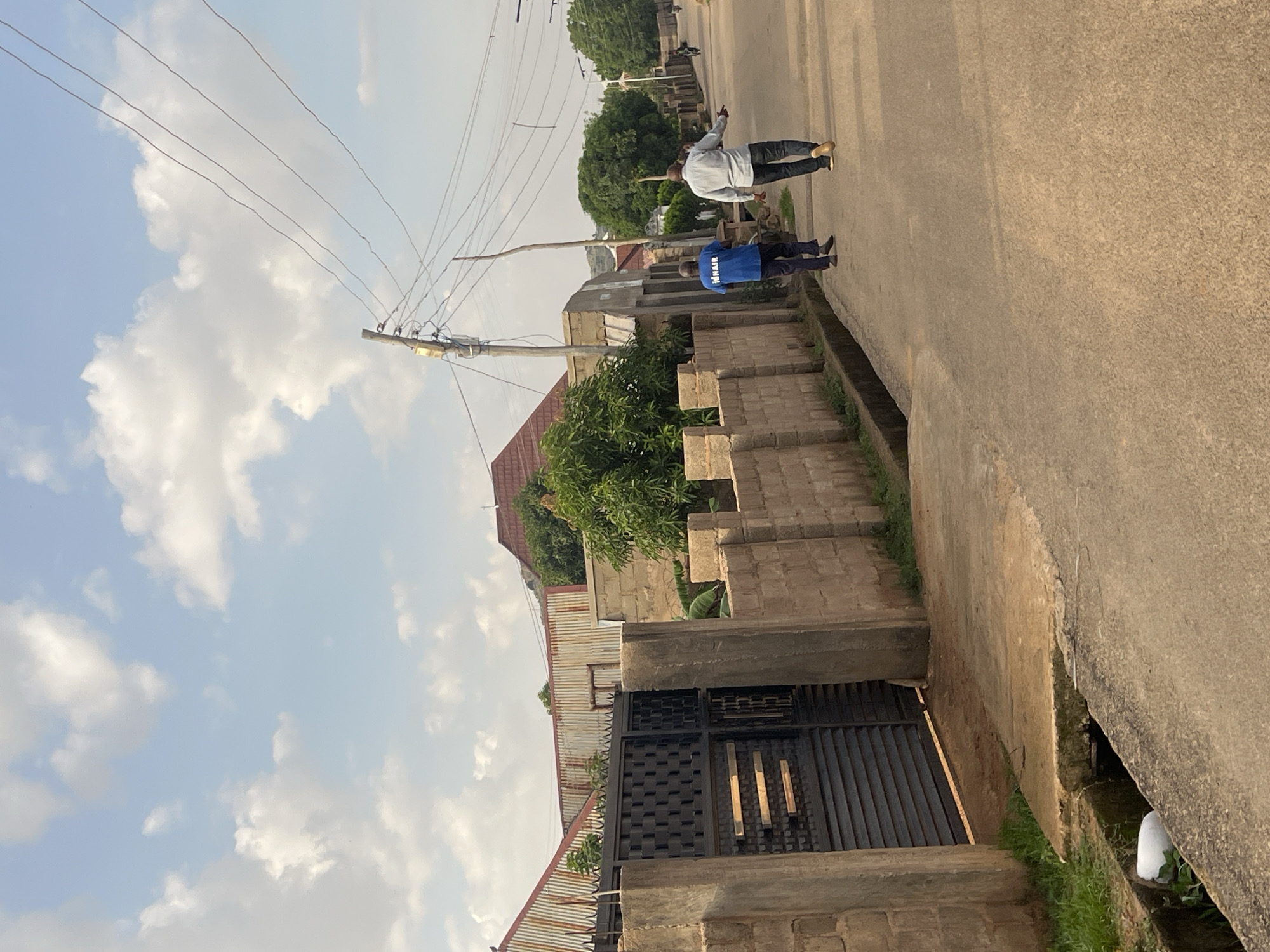 Two men are walking away from the camera down a paved street in the city of Jos, Nigeria. The man on the left is wearing a blue t-shirt with white lettering that reads "IDNAIR" and dark pants. The man on the right is wearing a light blue button-down shirt and dark pants. They are walking on the right side of the street.
To the left of the street, there is a low, light brown brick wall topped with a black metal gate and fence. Beyond the wall, there are buildings with corrugated metal roofs and a lush green tree. The sky is blue with scattered white clouds.
To the right of the street, there is a row of buildings and trees. There are utility poles and power lines running overhead. The street has a drainage ditch running along the left side. A white plastic bag is visible in the ditch. The overall scene depicts a typical urban streetscape in Jos.