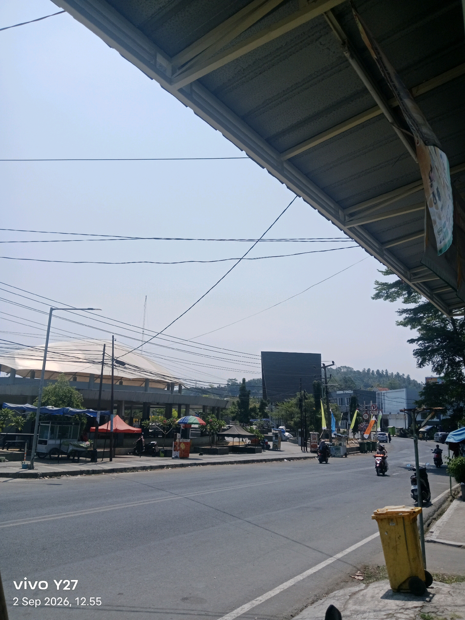 The image captures a street scene in Palabuhanratu, Indonesia, under a clear blue sky. A wide asphalt road dominates the foreground, with a white line marking the center. On the right side of the road, a yellow trash bin on wheels is prominent. Further down the street, several motorcycles are visible, with some riders in motion. On the left side of the road, there are various stalls and small buildings, some with awnings and umbrellas. A large, modern building with a curved roof structure is in the background, possibly a stadium or convention center. Power lines crisscross the sky. The image has a timestamp in the lower left corner indicating "vivo Y27 2 Sep 2026, 12.55".