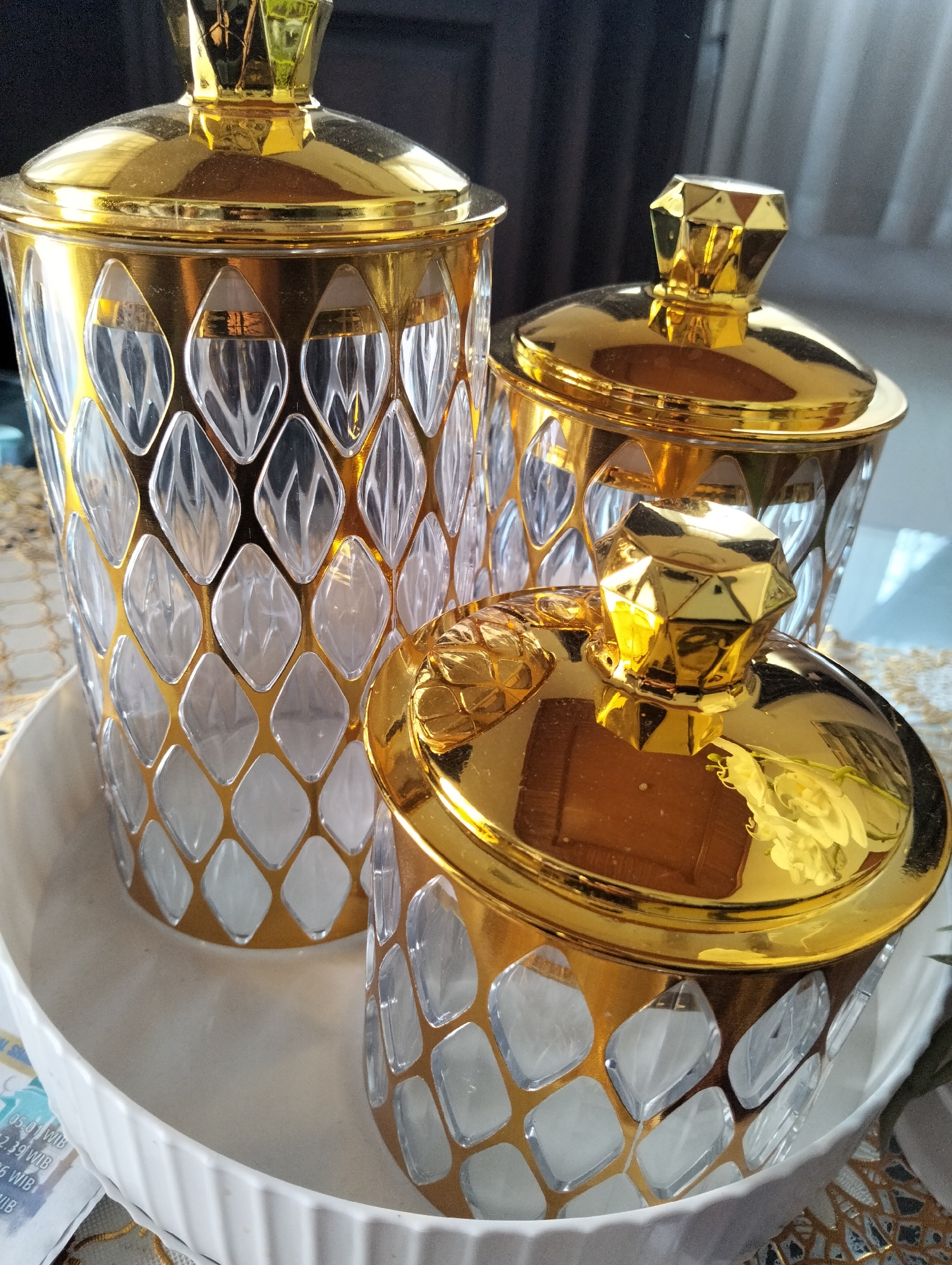 The image depicts three ornate, gold-accented jars with diamond-shaped glass panels, arranged in a white ceramic bowl. The jars are of varying heights and are positioned in a diagonal line, with the tallest jar at the back left, a medium-sized jar behind and to the right, and the smallest jar in the front right. Each jar has a matching gold lid topped with a faceted, jewel-like knob. The background consists of a dark curtain and a blurred window with natural light. A patterned tablecloth is visible beneath the bowl and jars. The scene appears to be an indoor setting, possibly a home or a decorative display. No people or actions are depicted. The location specified, Julicotmeurak, Indonesia, cannot be confirmed or denied from the visual information in the image alone.