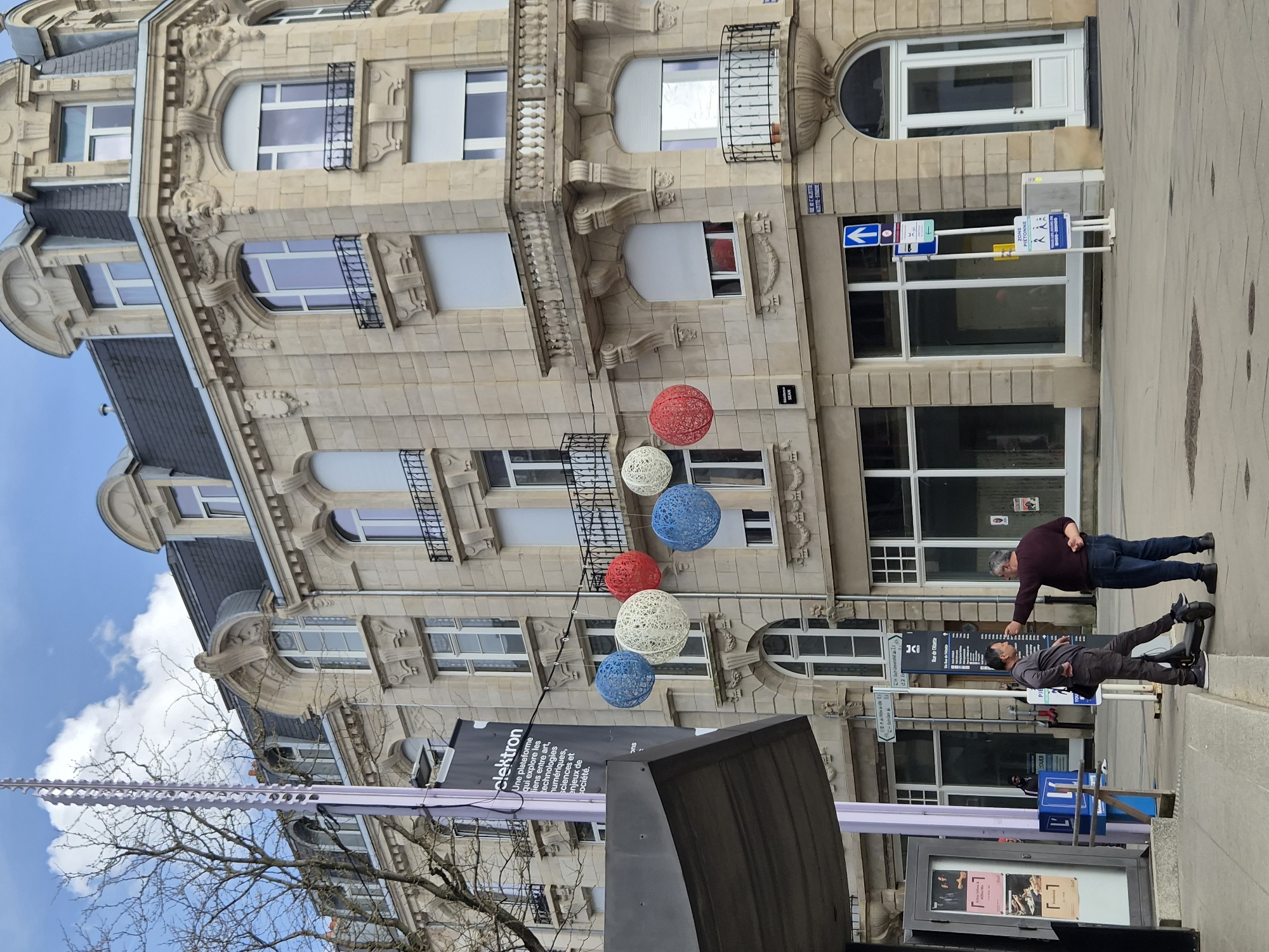 This image captures a vibrant daytime street scene in Esch-sur-Alzette, Luxembourg, showcasing classic European architecture and urban life.
The central focus is a grand multi-story building with a light-colored stone facade, adorned with intricate carvings, decorative balconies with wrought-iron railings, and arched window frames. The building features numerous large windows, some with exterior white roller blinds, and a dark-tiled roof with dormer windows.
Above the street, several large, spherical decorative lanterns made of woven material are suspended, colored in red, white, and blue, adding a festive touch.
On the paved sidewalk in the foreground, two men are interacting. One man, wearing a burgundy long-sleeved shirt and blue jeans, stands with his left arm outstretched, pointing his finger towards the other man. The second man, dressed in a grey top, dark trousers, and sneakers, is looking downwards, seemingly at something on the ground or in response to the first man's gesture.
Various street furniture and signs are visible. A prominent blue street sign points left with an arrow and reads "RUE DE L'ALLEE ALLEE-STR." and a smaller sign below it says "SEAN". Another yellow sign designates a "ZONE PIETONNE" (pedestrian zone) with pictograms. A large black banner hangs vertically, partially obscuring the building, with the text "clektron Une plateforme qui explore les liens entre art, technologies numériques, sciences et enjeux de société." A white street kiosk with posters inside and a sign "JCB" are also visible.
The weather appears clear with a bright blue sky and some white clouds, suggesting it's daytime. Bare tree branches are visible on the left, indicating either late winter or early spring.