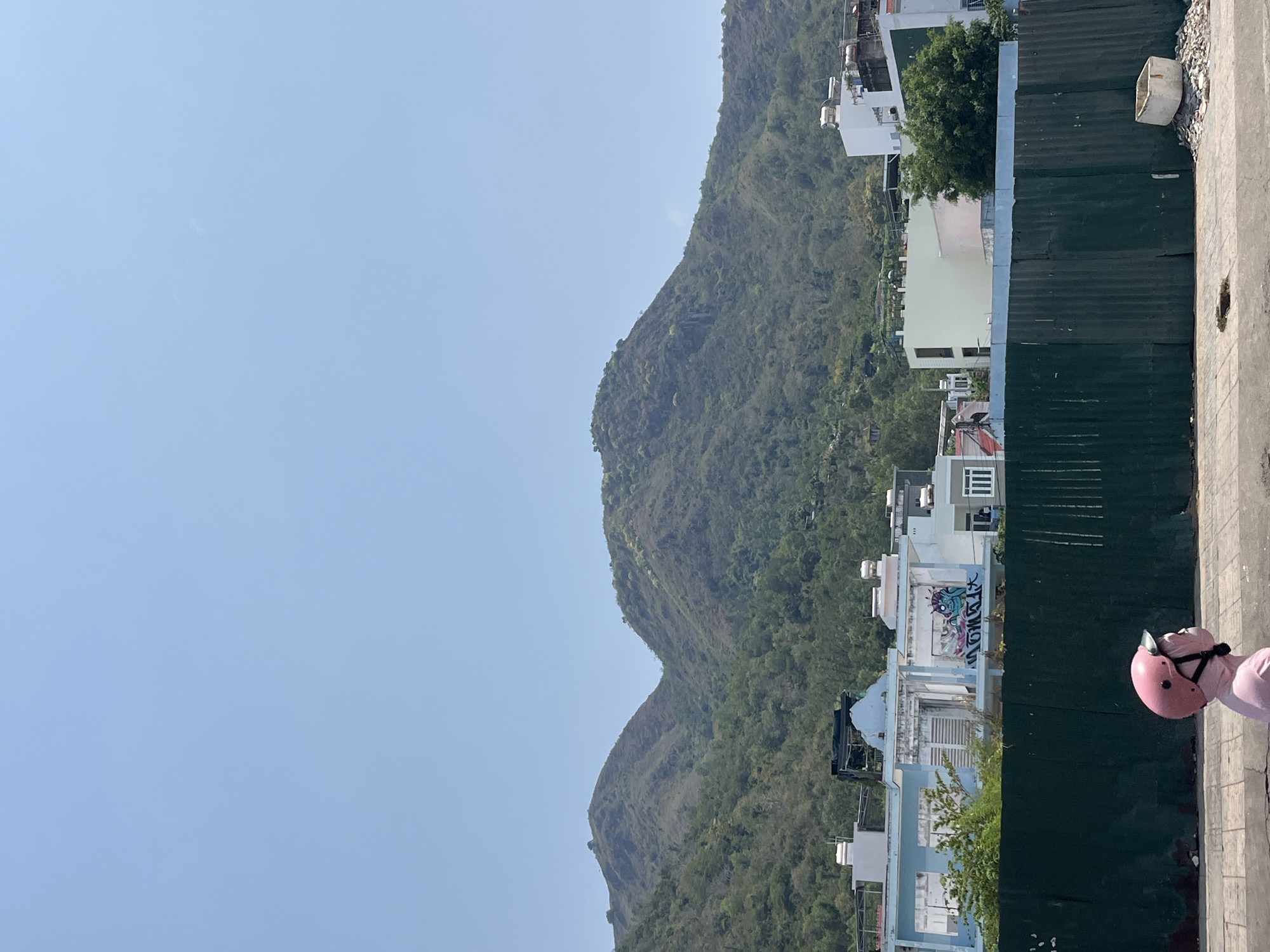 The image presents a view in or around Bac Nha Trang Ward, Vietnam. A mountain range covered in dense greenery occupies the central and right sides of the frame. Residential buildings, predominantly white or light-colored, are nestled along the lower slopes of the mountains. A green corrugated metal fence runs vertically along the right side of the image. A pink helmet rests on a cracked concrete surface in the bottom right corner. The sky above the mountain is clear and light blue.