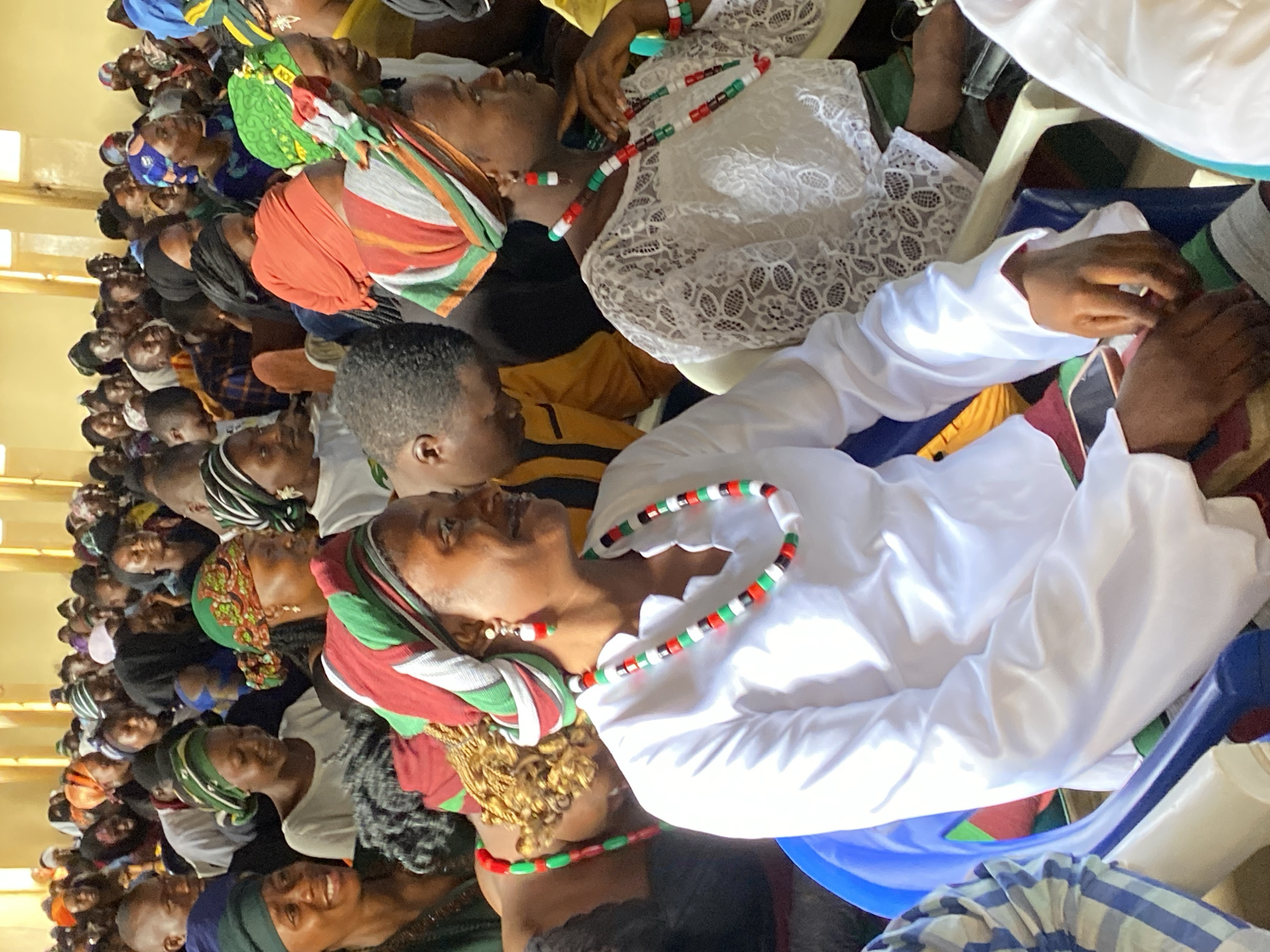 A dense crowd of people is seated indoors. Many individuals are wearing traditional African attire, including colorful headwraps and beaded necklaces in green, red, and white. One person wears a white lace dress and another wears a long-sleeved white shirt. The scene takes place indoors, likely in a hall or meeting space, with light-colored walls. The location is potentially Durimi, Nigeria.