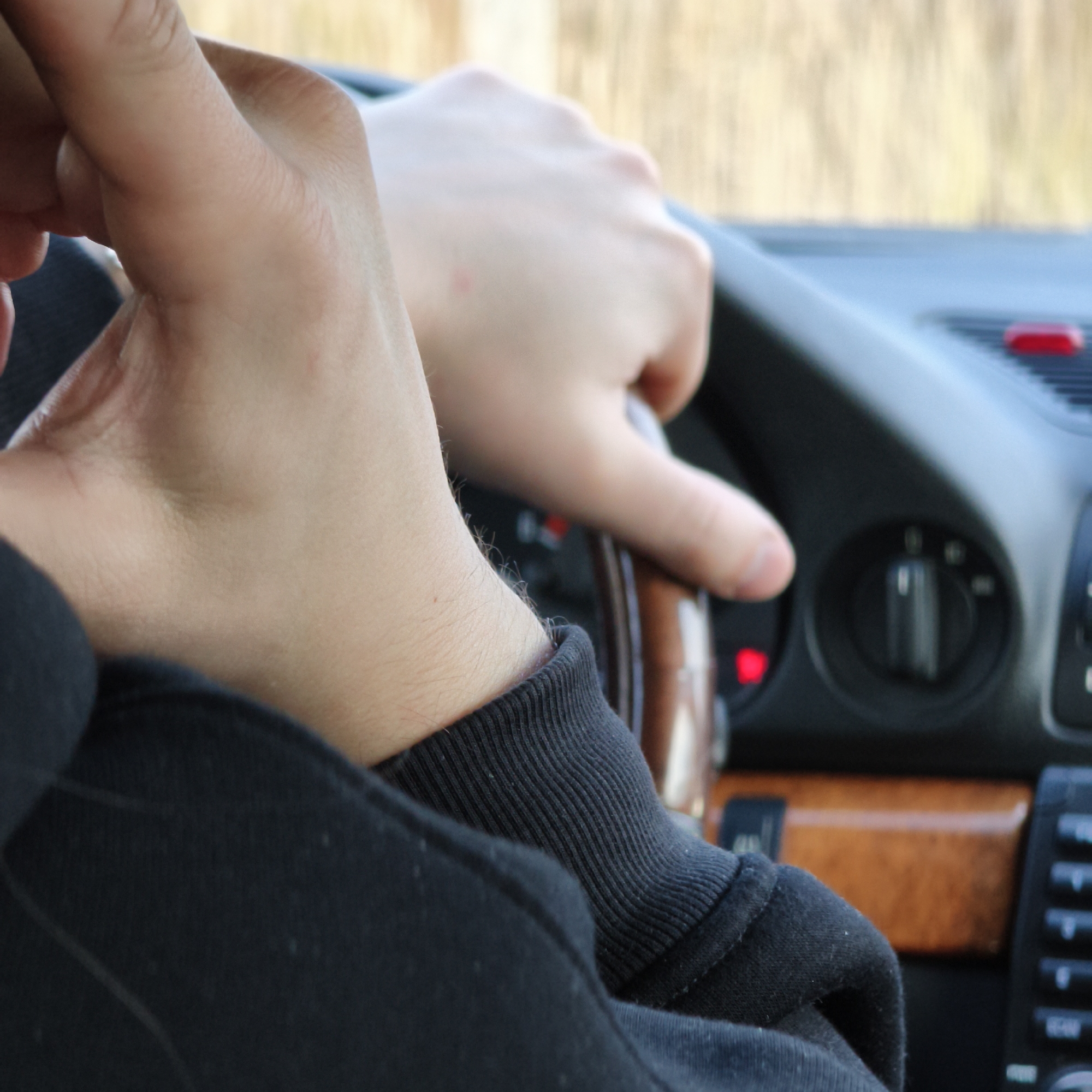 The image depicts a partial interior view of a vehicle, ostensibly in or near the city of Парэчча, Belarus. The hands and forearms of an individual are prominent. The right hand firmly grips the upper-right portion of a multi-material steering wheel, which features a dark, possibly wood-grain, section. The left forearm is partially visible, covered by a black long-sleeved garment with a ribbed cuff. The dashboard is visible in the mid-ground, featuring dark air vents, multiple control buttons, and at least two illuminated red indicator lights. A horizontal strip of polished wood trim is present on the dashboard. The background, visible through the windshield, is a highly blurred outdoor environment composed of light brown and yellowish hues, consistent with natural foliage or dry vegetation.
