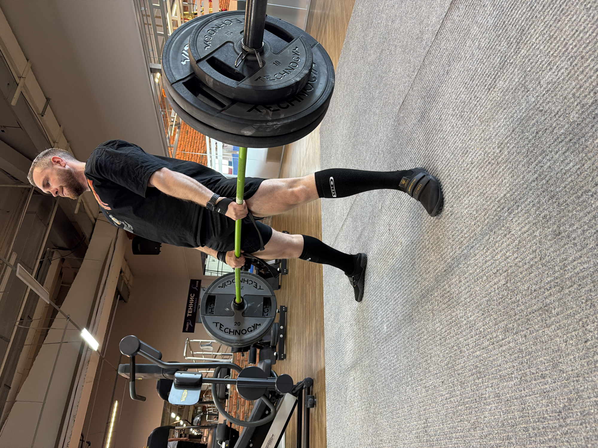 The image shows a man actively engaged in weightlifting within an indoor gym setting in Chaikovsky, Russia. The man, who has a beard and short hair, is dressed in black athletic wear, including a t-shirt, shorts, black knee-high socks with the "GILES" logo, black lifting gloves, and black minimalist training shoes branded "ARMSTRONG FITNESS."
He grips a barbell featuring a bright green center shaft and multiple black "TECHNOGYM" weight plates. Visible denominations on the plates include "20" and "10," with "4//" also visible on one plate. The man's head is tilted slightly down, and his focused expression suggests concentration during the exercise.
The gym environment includes a textured light grey carpeted floor transitioning to a wooden floor section. The ceiling reveals an industrial design with exposed structures and bright linear fluorescent lights. In the background, other gym equipment is partially visible, and a sign in Russian reads "ПОДАЧА ТЕННИС & SON" (Serve Tennis & Son), indicating a multi-sport or fitness complex. Another partially visible sign also displays "TECHNOGYM." The scene depicts a moment of intense physical training during an indeterminate indoor time of day.
