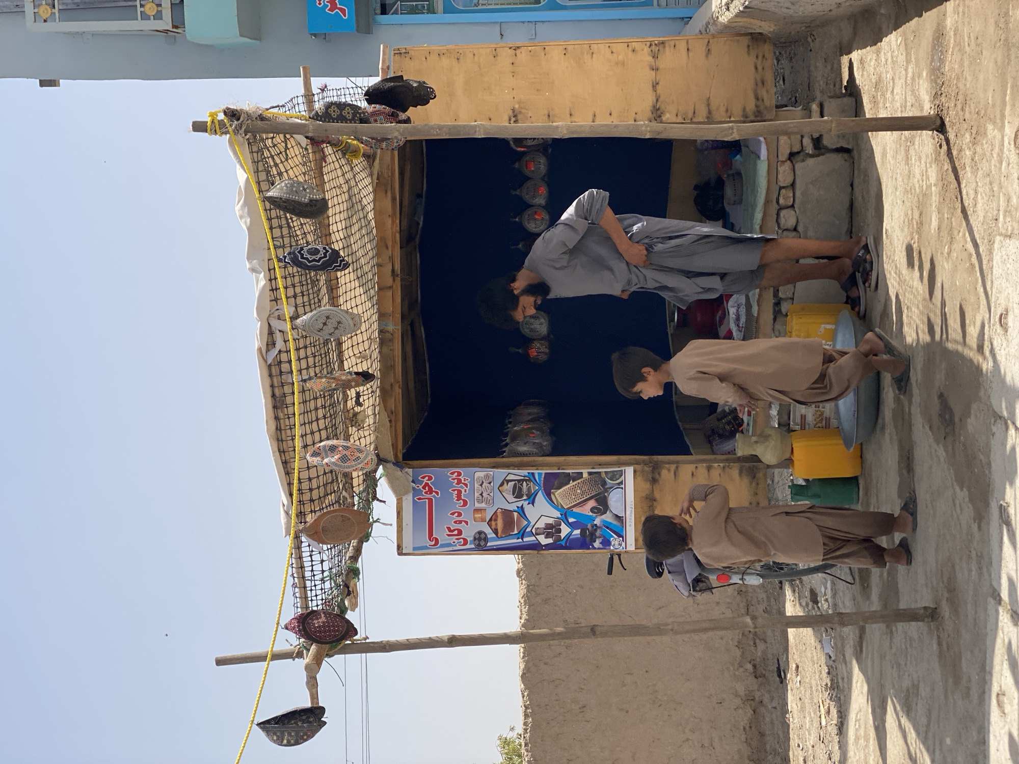 A man and two young boys stand outside a small, open-air shop in Kandahar, Afghanistan. The shop is constructed from wood and appears to be a temporary structure. It has a thatched roof covered with a net from which several embroidered caps are hanging. A banner with text in Arabic script and images of various items is displayed on the front of the shop. The man, dressed in a gray tunic and trousers, is standing in the doorway of the shop, looking down. One boy, wearing a light brown tunic and trousers, stands in front of the shop, facing away from the camera. The second boy, also in similar attire, stands to the right of the first boy, also facing away. In the background, a motorcycle is parked next to the shop. Yellow and green plastic containers and a metal basin are visible on the ground near the entrance. The environment appears to be dusty and dry, with a concrete walkway in the foreground. The sky is clear and blue.