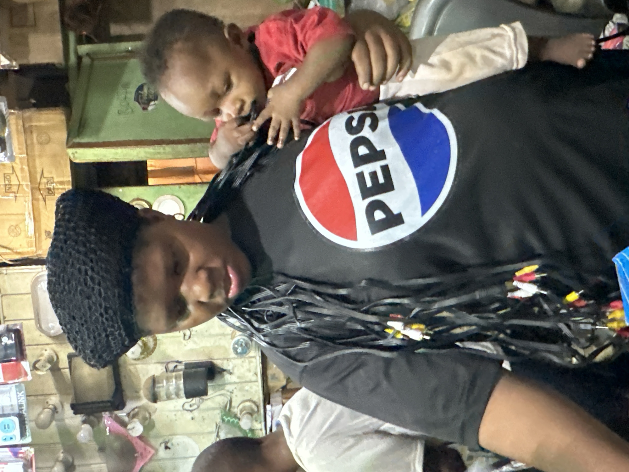 In Narayi, Nigeria, a woman wearing a black knitted cap and a black t-shirt with a Pepsi logo stands with a baby on her left shoulder. The baby, dressed in a red shirt and light-colored pants, is being held by the woman's right arm and is chewing on something. The woman has black braided hair that is adorned with colorful RCA connectors. Behind the woman, a man in a white t-shirt is partially visible. The background appears to be an indoor space with shelves holding various items, including boxes and what looks like lighting fixtures. A green cabinet is also visible on the right side of the image.