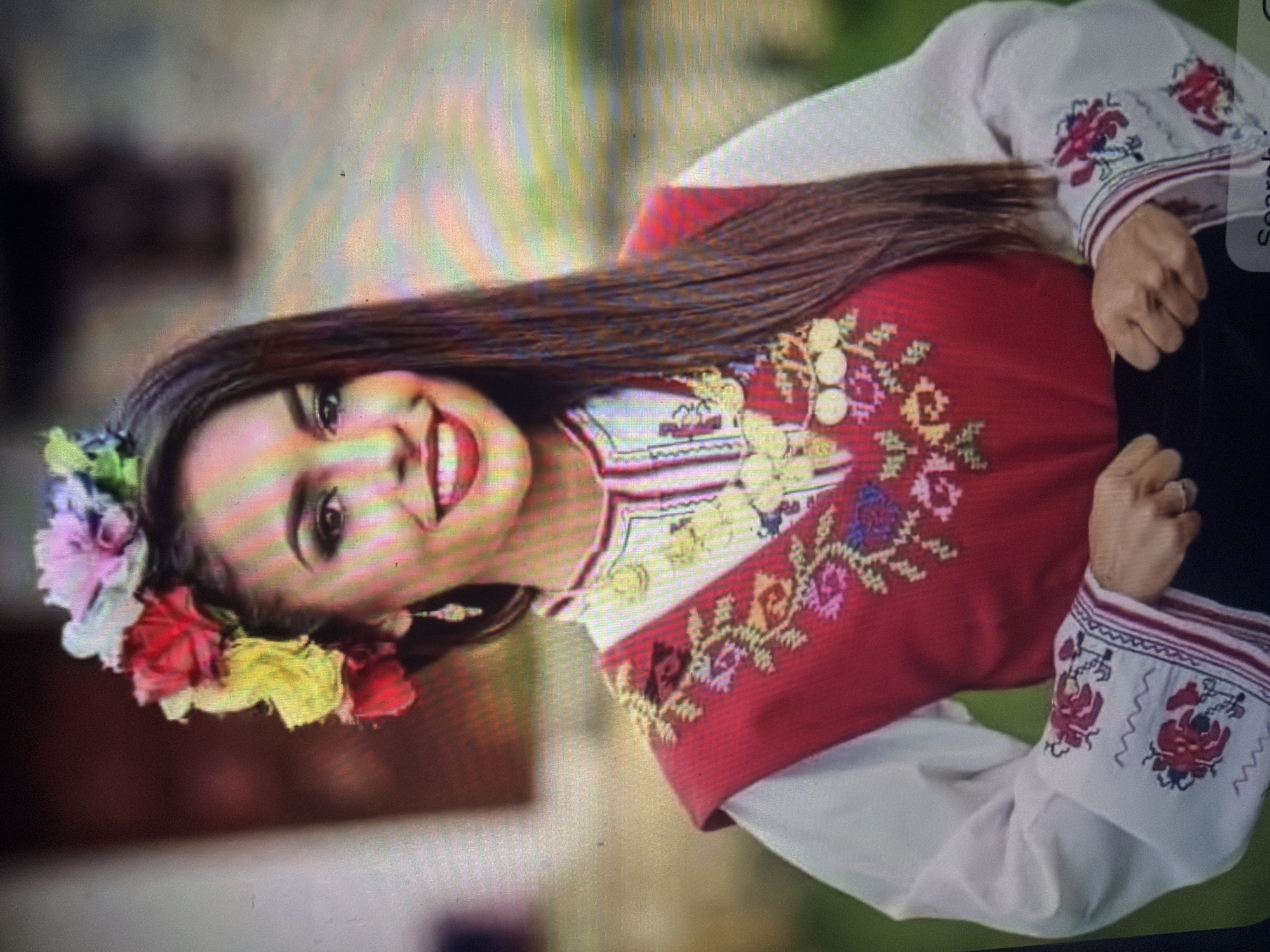 A young woman is depicted in a medium shot, smiling and looking directly at the camera. Her dark hair is long and straight, parted on the side, and adorned with a vibrant floral crown featuring red, yellow, pink, purple, and green blossoms. She wears dark eye makeup and red lipstick, with dangling earrings visible. The scene is situated in Shumen, Bulgaria.
Her attire is a traditional Bulgarian folk costume. She wears a white long-sleeved blouse with intricate red, white, and dark blue embroidery on the cuffs and around the neckline. Over the blouse, she has a sleeveless red garment, likely a sukman or sarafan, which is heavily embroidered with multi-colored patterns including yellow, green, pink, purple, blue, and red floral and geometric motifs on the chest and shoulder areas. A large, multi-strand gold-colored necklace, featuring circular coin-like pendants, is prominently displayed over the red garment. Her hands are clasped together at waist level, and a ring is visible on her right hand. The background is softly blurred, showing green foliage on the right and a lighter, out-of-focus architectural structure on the left, indicating an outdoor setting under natural light.
