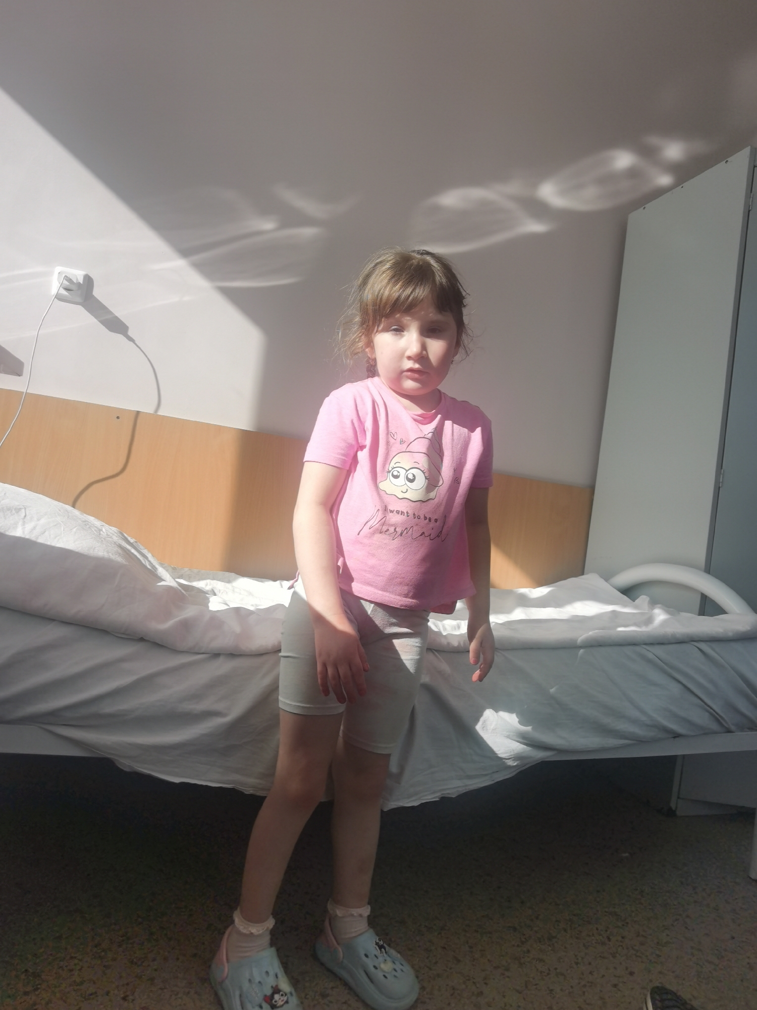 A young child stands in a room, possibly a hospital room, near a bed. The bed has a white sheet and a light wooden headboard. The child is wearing a pink t-shirt with a cartoon graphic, gray shorts, white socks with lace trim, and blue clogs with a character design. A white wall is behind the bed, with a wall socket and light shadows cast upon it. The room appears to be brightly lit. The exact location in Perm, Russia, is not evident from the image.