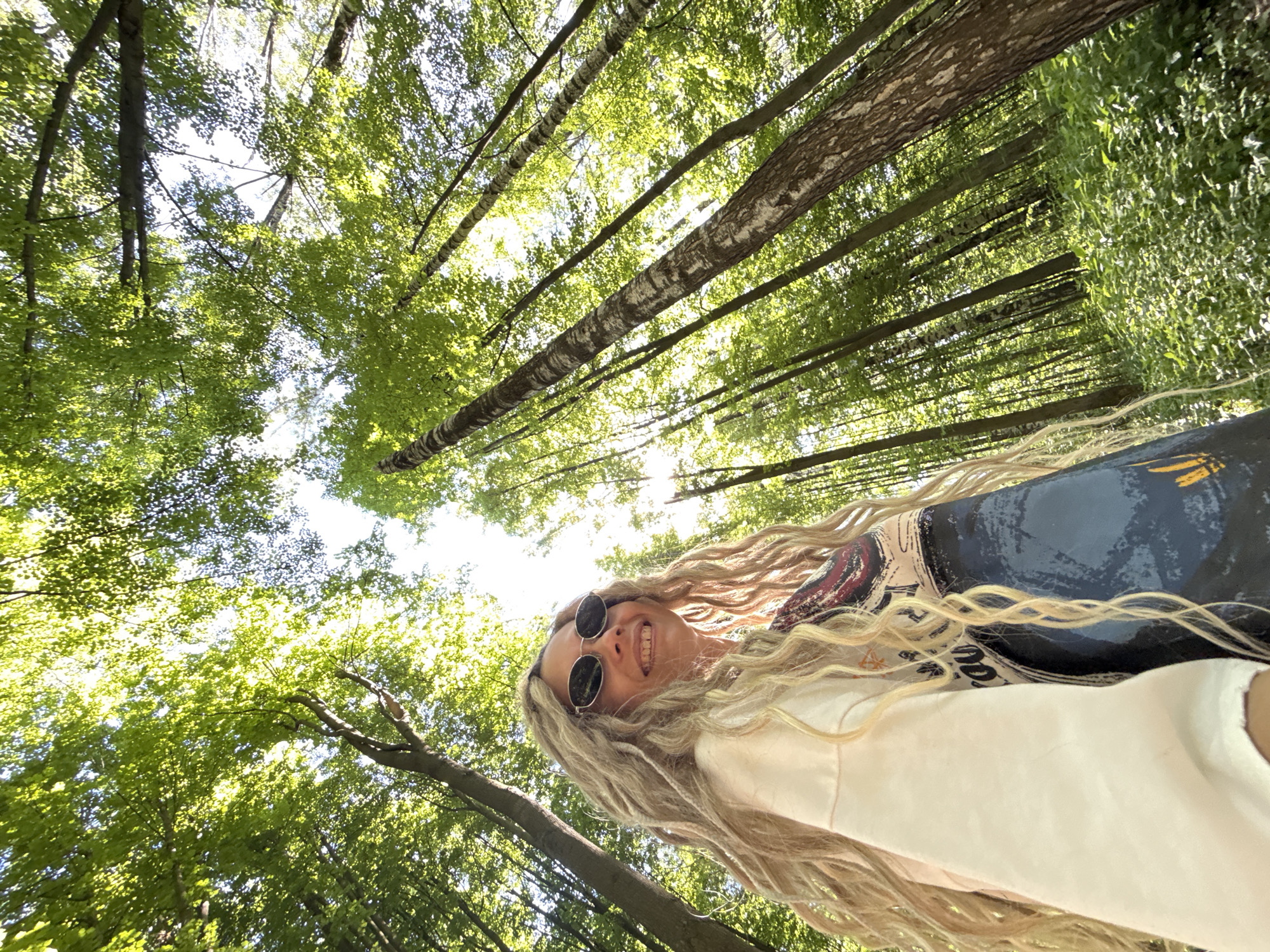The image displays a low-angle perspective within a forest, identified as being in Rusanovo, Russia. In the foreground, positioned in the lower left, is an individual with long, blonde, textured hair that appears as dreadlocks. This person is wearing dark, oval-shaped sunglasses and a light-colored long-sleeved shirt over a patterned darker garment. Their head is tilted slightly upwards and to the right, revealing a smiling expression. The environment is characterized by a dense forest composed of numerous tall, slender trees with vibrant green foliage, indicative of a deciduous forest in full leaf. Strong sunlight filters through the canopy, creating bright highlights among the leaves and illuminating the upper sections of the tree trunks. Lush green undergrowth covers the forest floor visible in the lower right quadrant of the image. The camera angle emphasizes the height of the trees and the expansive canopy.