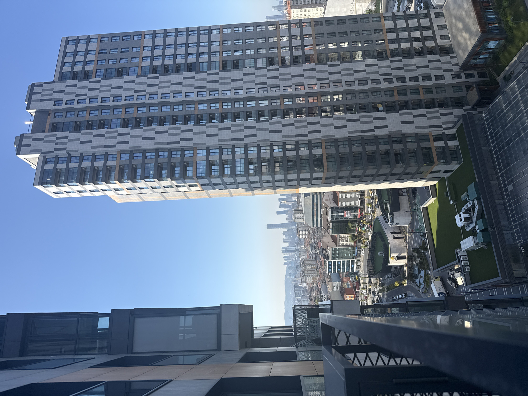 The image is an elevated, exterior, eye-level view of a modern cityscape in Ümraniye, Turkey, taken on a clear day with a bright blue sky. In the foreground, a portion of a dark-colored balcony railing is visible on the left, with a building facade constructed of wood-toned panels and dark-tinted windows on the left edge of the frame. To the right of this building is a very tall, modern skyscraper with a distinctive gray and white patterned facade, featuring numerous balconies and rows of dark-framed windows.
Further in the background, a dense urban landscape unfolds, characterized by a variety of buildings of different heights and architectural styles, many with flat roofs. A distant skyline of taller skyscrapers is visible on the horizon, suggesting a significant urban center. Below the main tall building, a complex with green rooftop areas and what appears to be a McDonald's restaurant with a circular design and water feature is observable. Streets with moving vehicles are also visible in the middle ground. The overall impression is of a contemporary, developing urban environment.