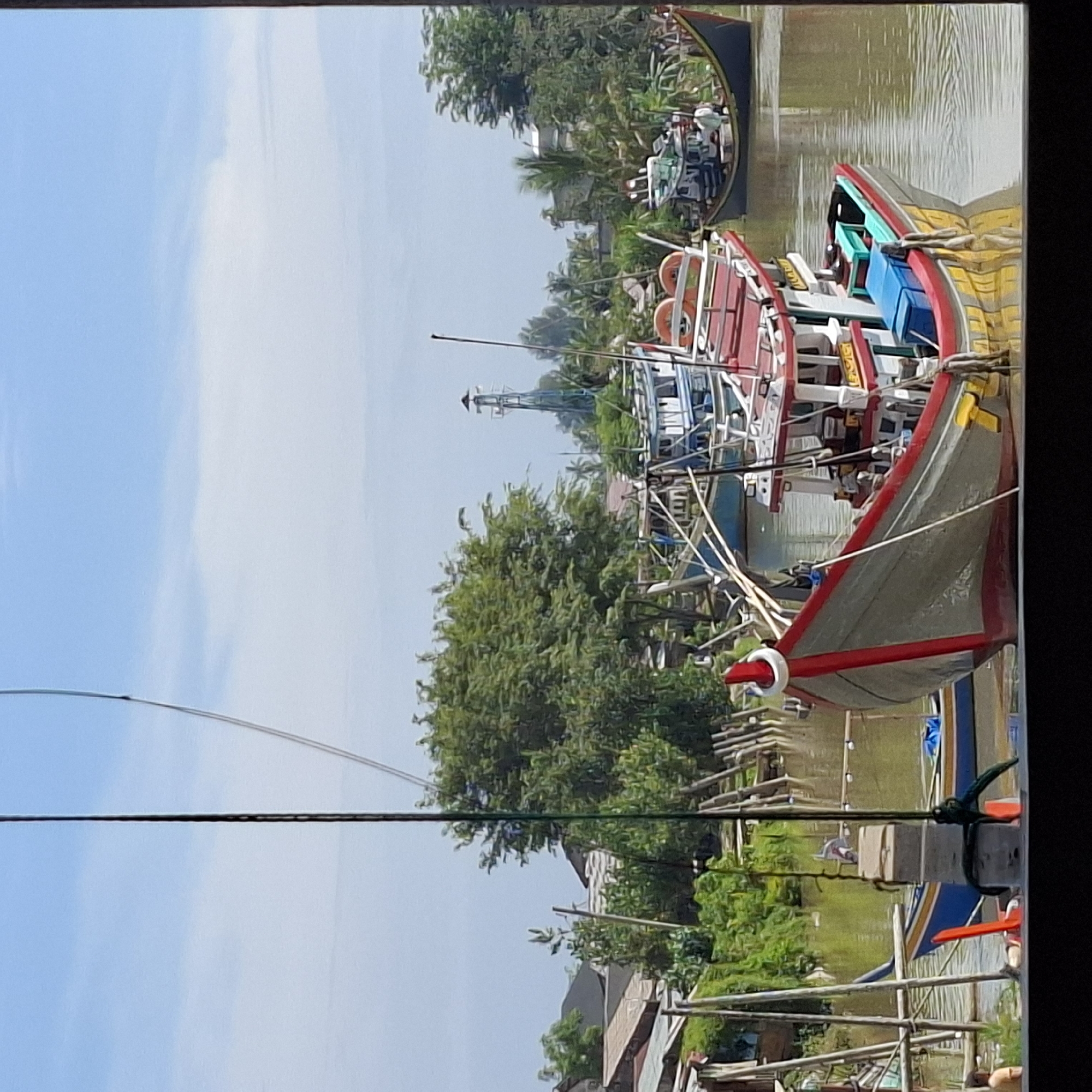The image depicts a waterfront scene in the city of Mesjid Tuha, Indonesia. The foreground is dominated by the bow of a gray and red fishing boat, its hull brightly painted with yellow and red stripes, and equipped with blue storage boxes. The boat is moored in a brown river. Behind it, another fishing boat, primarily white with blue trim, is visible. Further in the background, a variety of other boats are present, as are lush green trees and some houses with corrugated roofs, indicative of a residential area along the water. A tall, blue, lattice-like structure, possibly a lighthouse or communication tower, stands on the horizon amidst the trees. The sky is a pale blue with scattered white clouds. The presence of wooden posts and bamboo structures along the riverbank suggests a functional dock or pier area. Two individuals are partially visible on the left side of the image, one in an orange shirt and another sitting down. The overall impression is one of a working harbor or a settlement situated on a river.