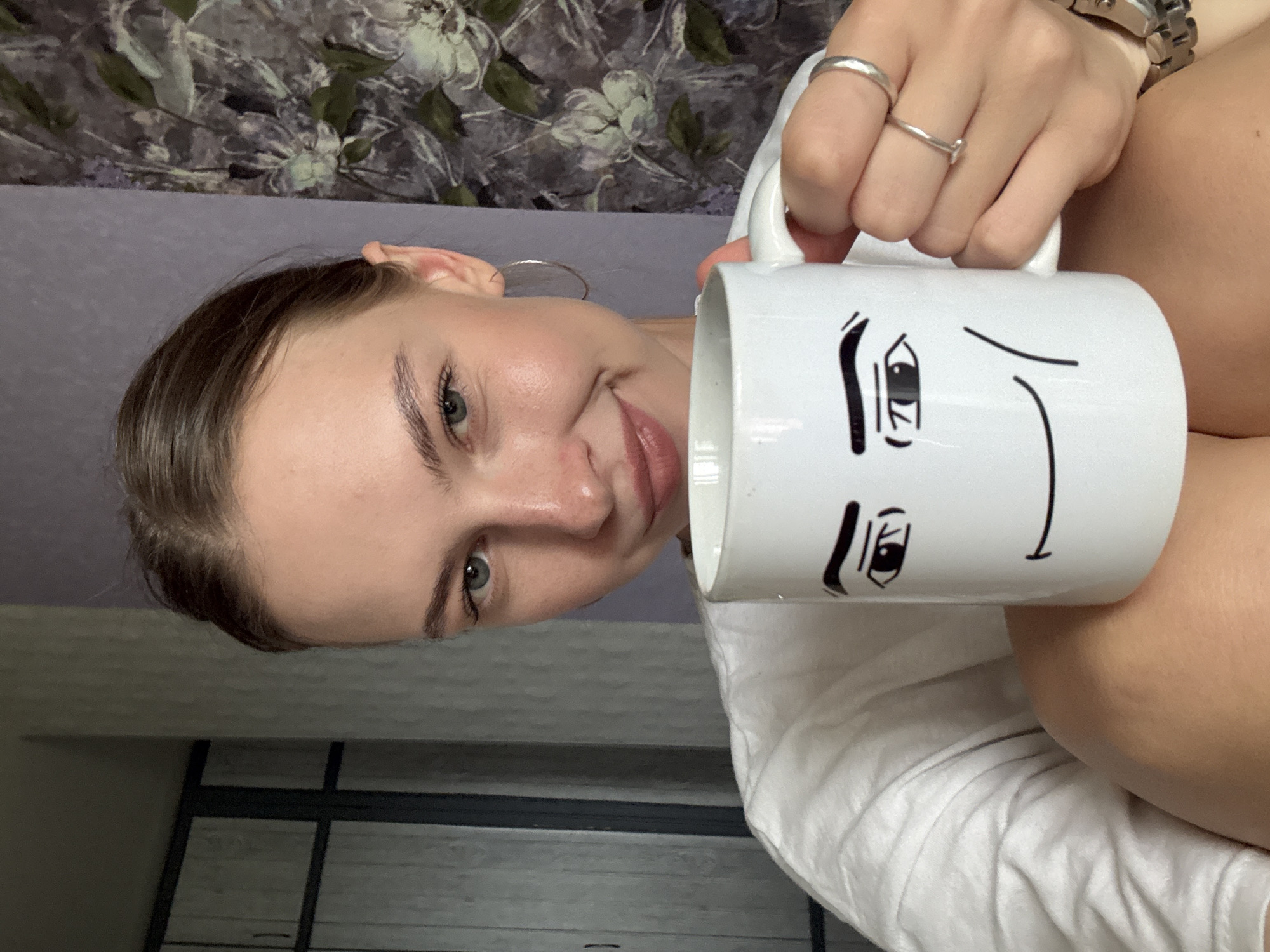 A young Caucasian woman is holding a white mug with a cartoon face drawn on it. She is wearing a white t-shirt and a silver ring on her left hand, and another on her right hand. She is also wearing hoop earrings. Her hair is pulled back, and she is smiling. The background appears to be an indoor setting with a floral wallpaper. The image is taken from chest up. The location is not identifiable as Perm, Russia.
