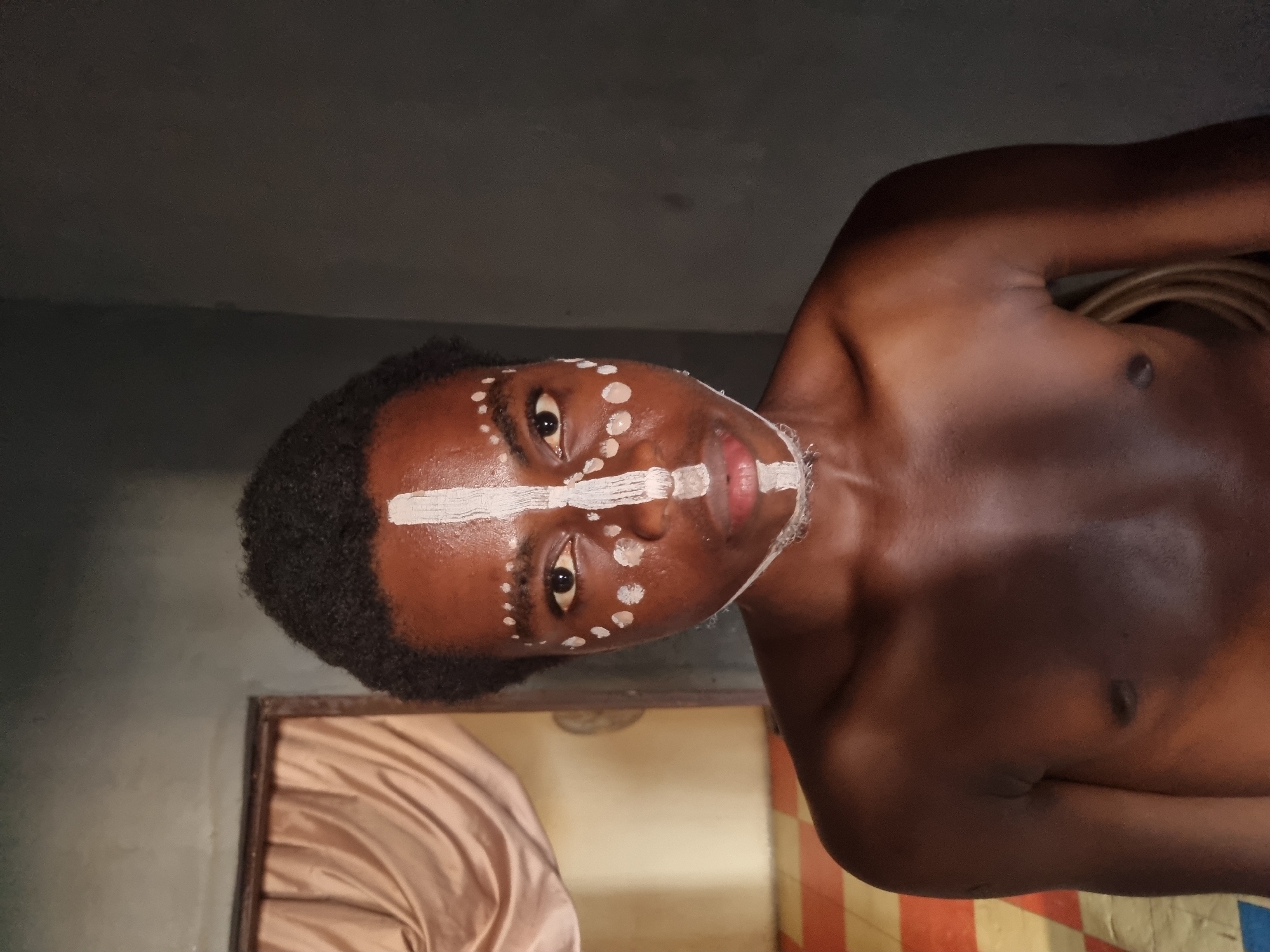 A dark-skinned person with short, dark hair is photographed indoors against a backdrop of a grey wall. The person is bare-chested and has white paint markings on their face, including a stripe across the forehead, a stripe across the nose, and dots on the forehead and cheeks. A doorway with a tan curtain is visible behind the person, and a striped wall can be partially seen on the right side. The photograph is taken at an angle. The specific location within Onitsha, Nigeria, cannot be determined from the image alone.