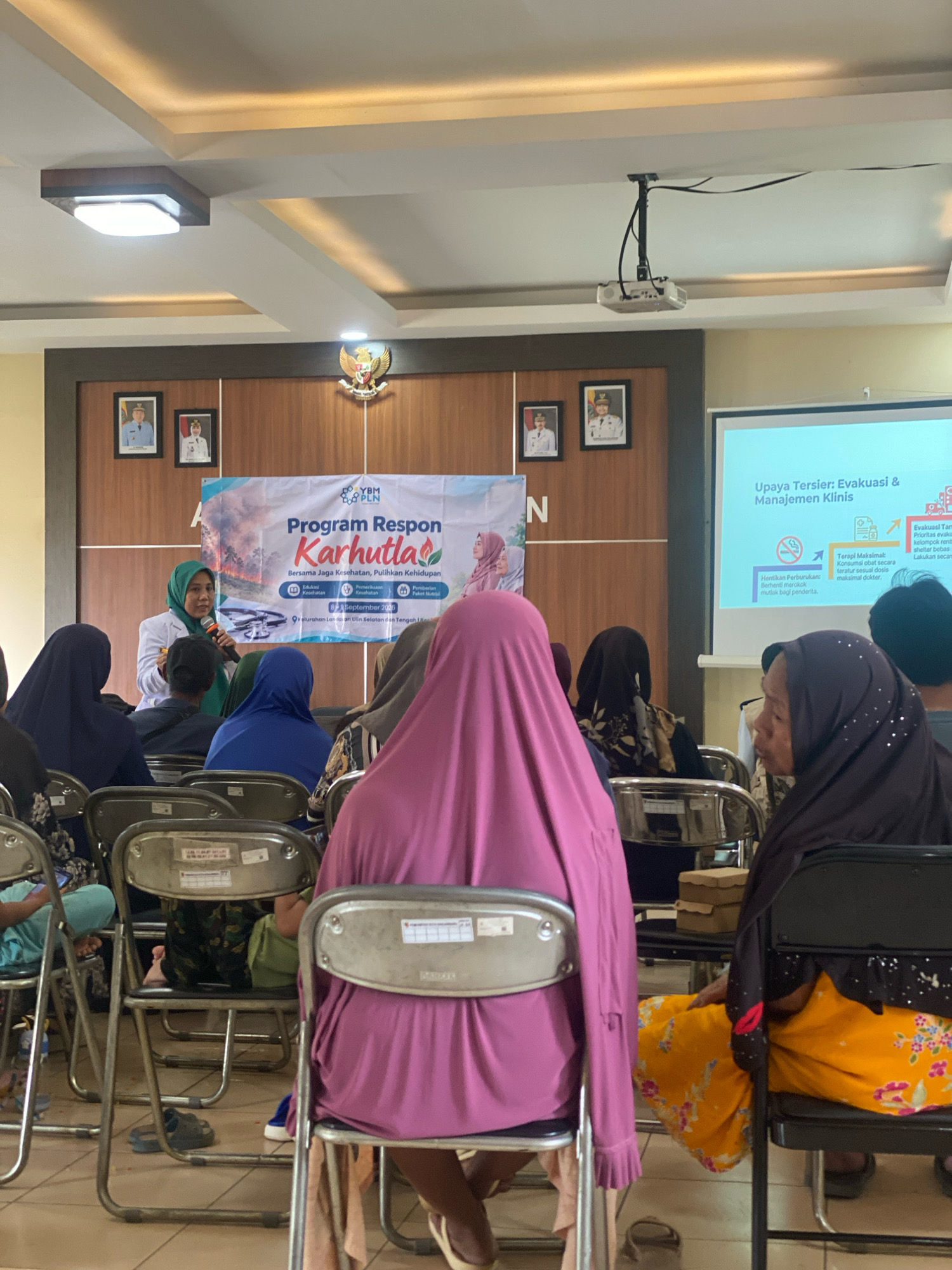 The image depicts an indoor event, likely a seminar or presentation, taking place in Banjarbaru, Indonesia. A woman wearing a white lab coat and a hijab is speaking into a microphone, addressing an audience seated in chairs. Behind her, a banner with text and images is displayed. To the right, a projection screen shows text in Indonesian, likely related to the presentation topic. The room has a wooden paneled wall and a ceiling with recessed lighting and a projector mounted overhead. Several framed portraits of men in official attire are visible on the wall. The audience members are predominantly women wearing hijabs in various colors. Some chairs are empty, and a few people are visible from behind. The flooring appears to be tiled. The atmosphere suggests a formal or semi-formal gathering focused on health or disaster response, given the banner's text "Program Respon Karhutla" (Forest and Land Fire Response Program) and the presentation screen's content.
