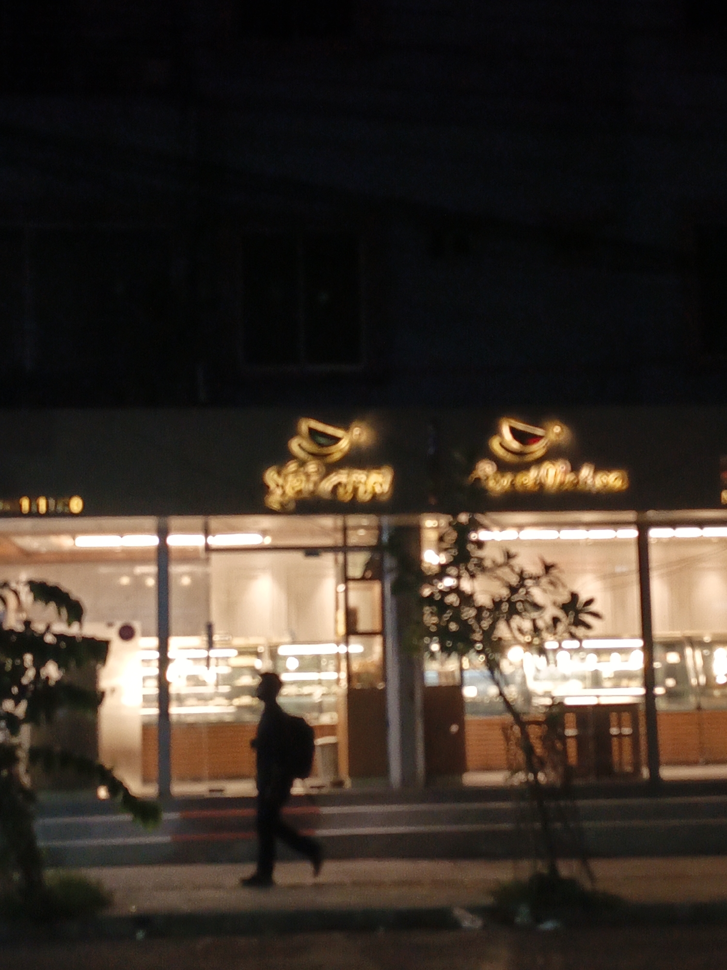 The image shows a storefront at night with bright interior lighting illuminating display cases. A person is walking along the sidewalk in front of the store. The store's signage includes a logo with a cup or bowl image and text reading "Amal Melaka." The city and country are unknown.