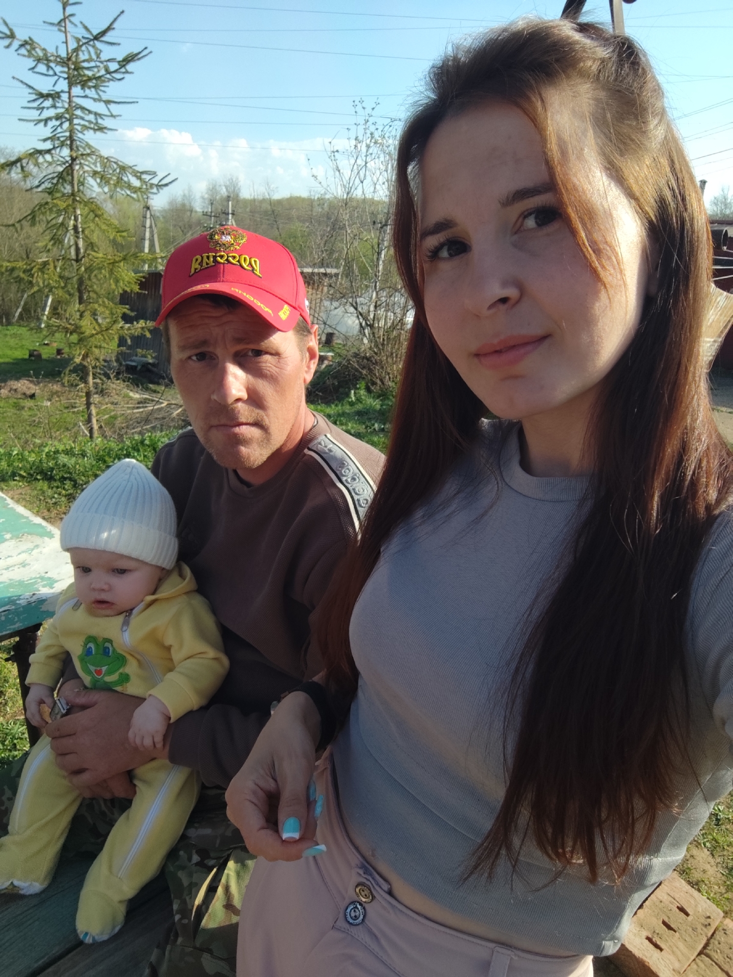 A photo captures three individuals outdoors in Фоки, Russia. A woman with long, brown hair and fair skin, wearing a light gray long-sleeved shirt and light pink pants with decorative buttons, is positioned on the right side of the frame, likely taking a selfie. Beside her and slightly behind, a man in a brown shirt and a red baseball cap holds a baby dressed in a yellow jumpsuit with a white knit hat. The baby's outfit features a green frog design on the chest. They are seated on what appears to be a bench or edge of a table painted green. The background shows a rural or suburban setting, with trees, grass, and elements of a home or structure visible. The sky is blue, indicating it is daytime.