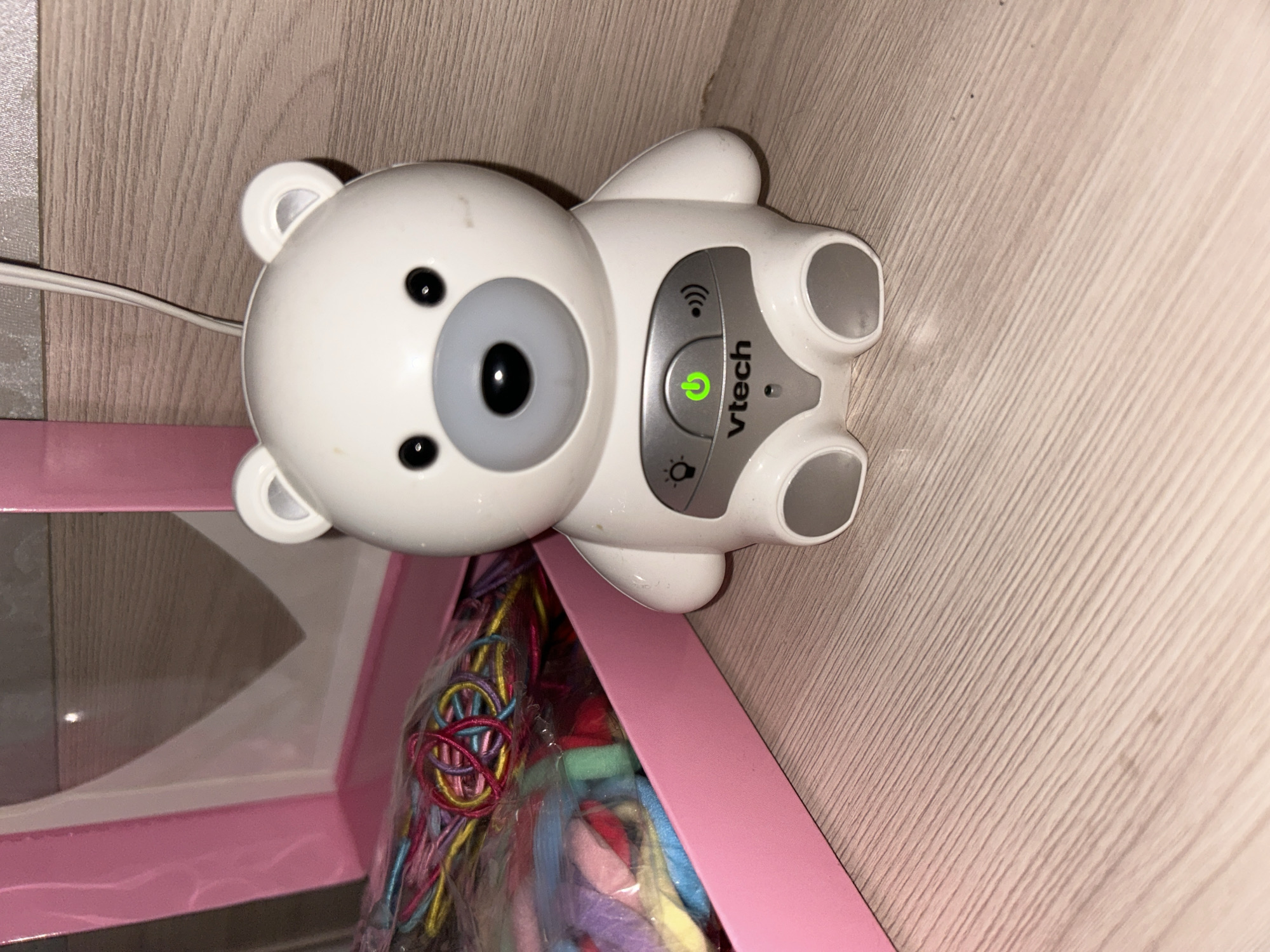 In Primorka, Russia, a white VTech bear-shaped baby monitor is positioned on a light wood-grained surface. The monitor features a grey snout, black eyes, and a green power button illuminated on its chest. To the left of the monitor is a pink box containing a bundle of colorful strings. The background consists of a plain, light-colored wall.