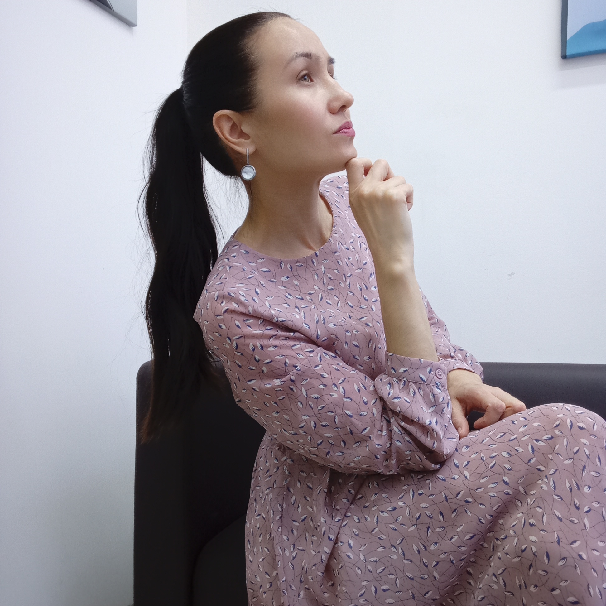 A woman with dark hair pulled back into a ponytail is sitting on a dark-colored couch. She is wearing a long-sleeved, mauve dress with a small leaf pattern. Her right hand is resting on her chin in a thoughtful pose. She is looking upwards and to her right. She is wearing a round, light-colored earring in her left ear. The background is a plain white wall with a framed picture visible in the upper right corner. The location is unspecified, and there is no indication of it being in Izhevsk, Russia.