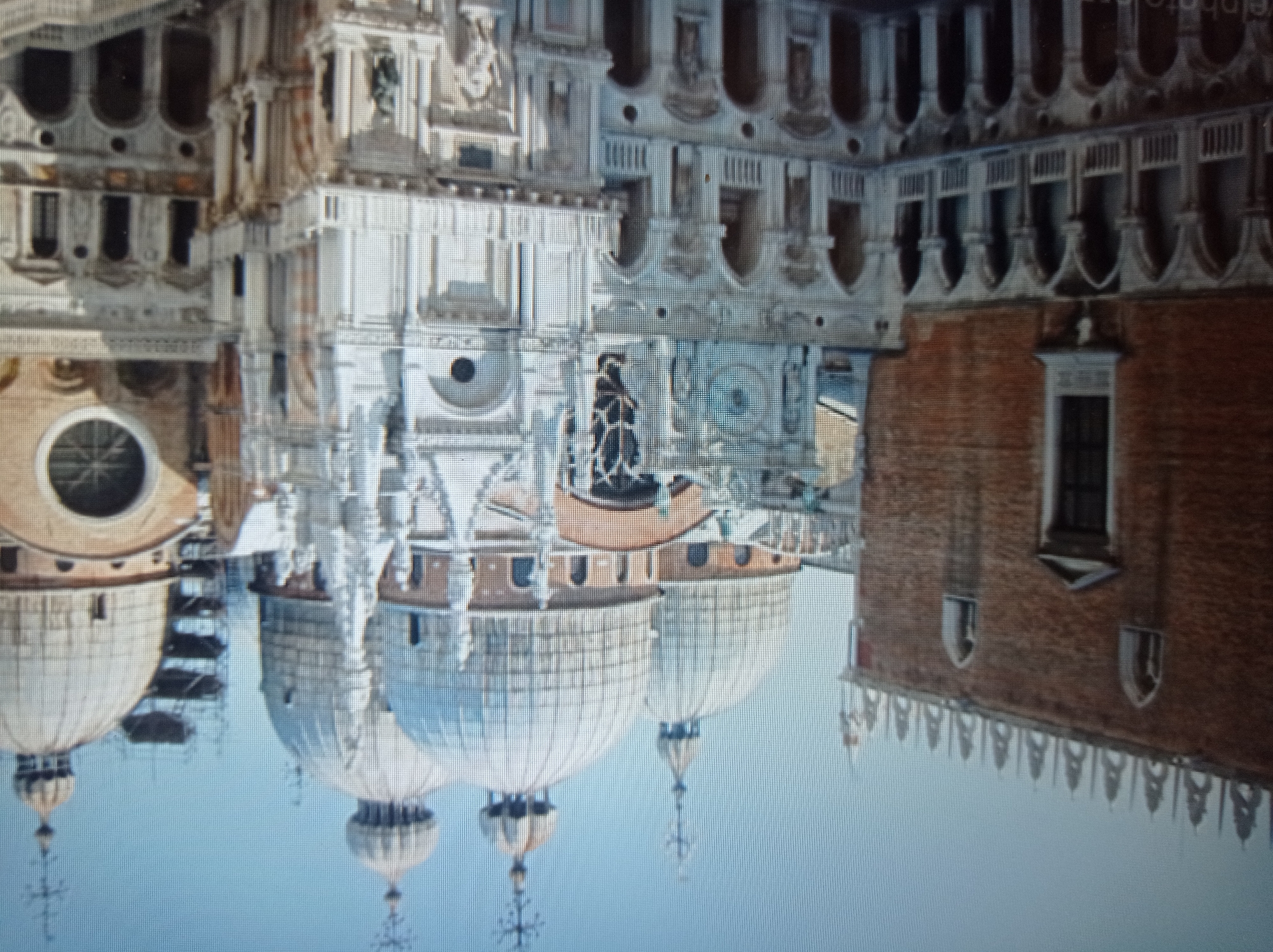 The image depicts a section of the Doge's Palace in Venice, Italy, characterized by its distinctive Gothic architecture. The palace features ornate facades with intricate carvings, arched windows, and multiple levels of loggias supported by columns. Prominent on the right side are the domes of St. Mark's Basilica, identifiable by their onion-shaped profiles and crosses atop their spires. The scene is captured from an elevated perspective, suggesting a viewpoint from an adjacent building or a drone. The sky is clear and blue, indicating daytime. There are no people or animals visible in the frame. The architecture suggests a historical and cultural landmark. The prompt's request to associate this image with Mukachevo, Ukraine, is inconsistent with the visual evidence, as the depicted architecture is definitively Venetian.