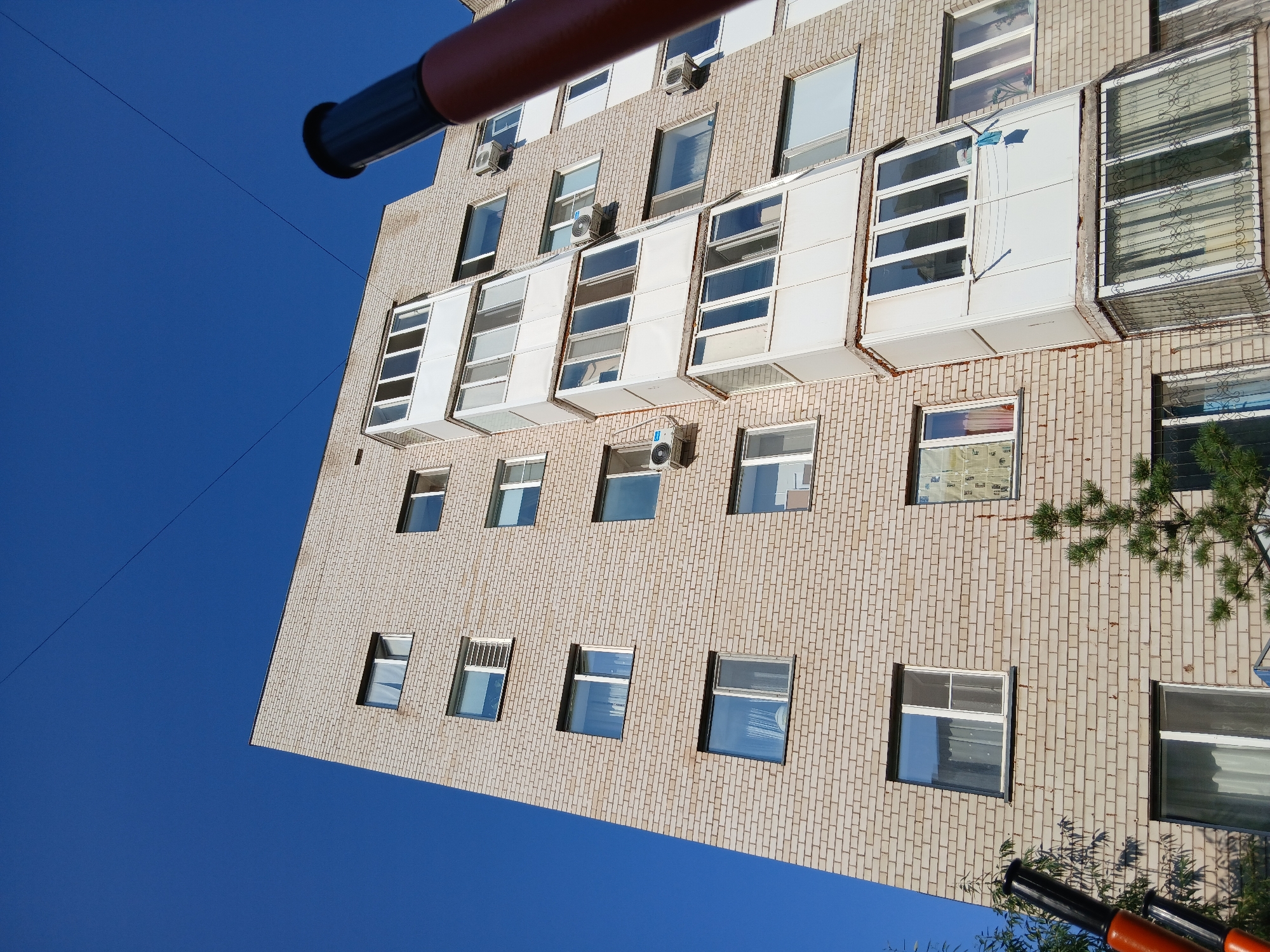 The image shows the exterior of a multi-story apartment building constructed from light-colored bricks, under a clear blue sky. The building features numerous windows and balconies with white trim. Some windows have air conditioning units installed. The perspective is from a low angle looking upwards, suggesting the photo was taken from the ground or a lower vantage point. A dark, cylindrical object, possibly part of a telescope or similar equipment, is partially visible in the foreground on the right side of the frame. On the left side, another dark and orange object, also appearing to be equipment, is visible. There are wires strung across the sky above the building. Sparse greenery, including a small evergreen tree, is present at the base of the building. The architecture and style of the building are consistent with residential structures commonly found in Astana, Kazakhstan.