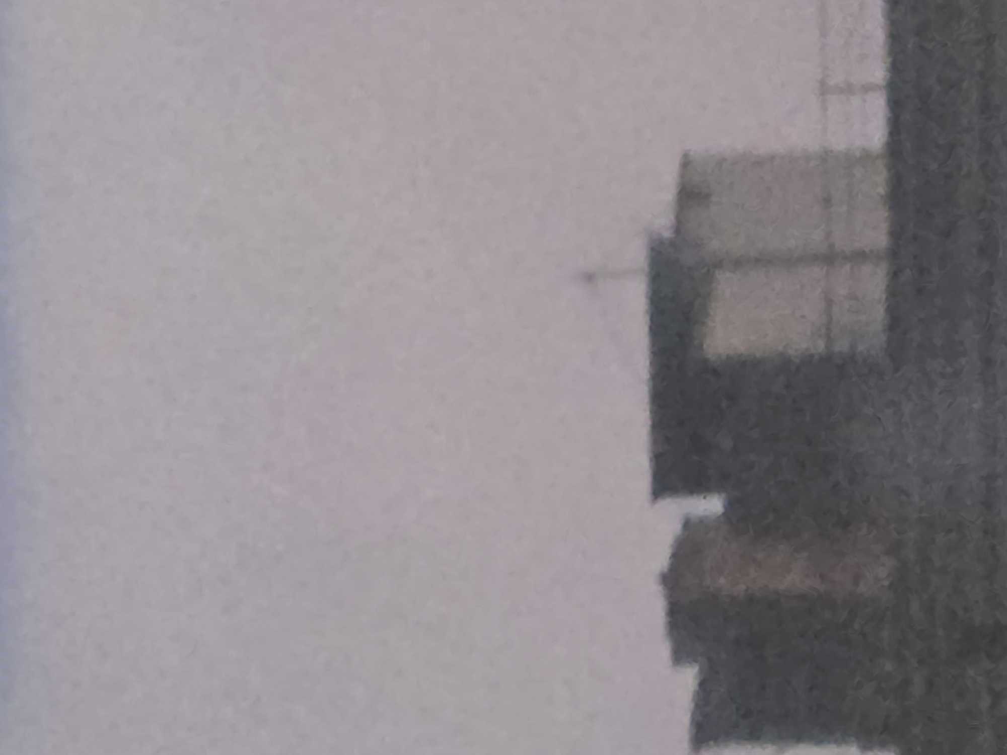 A distant, hazy, medium-shot photograph shows the tops of several buildings against a pale, overcast sky. The buildings appear to be on a rooftop. One building on the far left is partially visible, dark and rectangular. To its right, a taller, narrower building stands, a lighter tan color. Next to this, a dark, flat-topped structure is visible. To the right of this, a lighter-colored, boxy structure rises, with what appears to be a railing running horizontally across its lower section, suggesting a balcony or rooftop access. A thin, dark, vertical antenna-like object extends from the top of the center-right building. The overall impression is one of urban architecture seen from a distance through atmospheric haze.