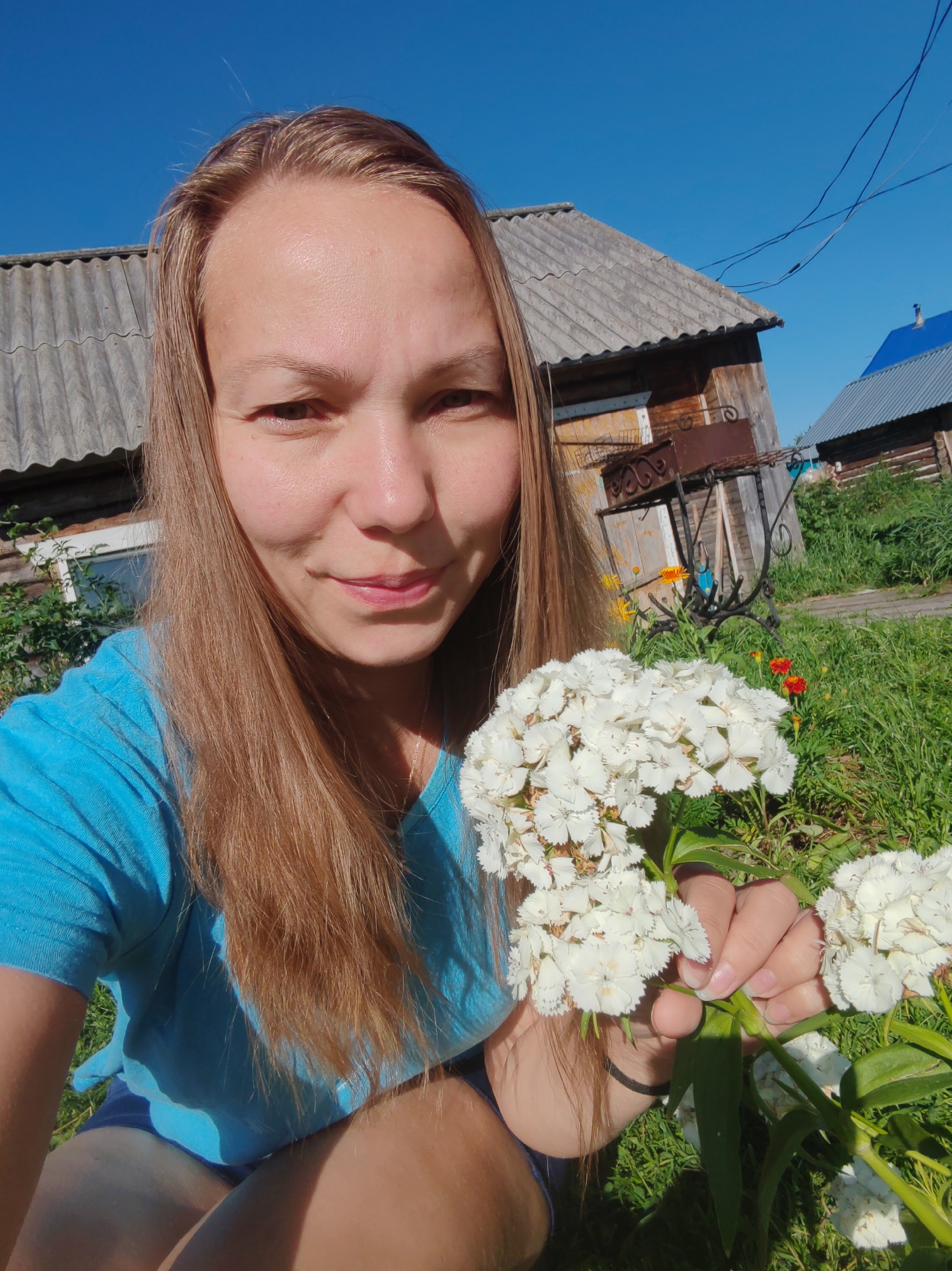 A woman with long, light brown hair is holding a bouquet of white flowers and taking a selfie outdoors. She is wearing a blue t-shirt and dark shorts. The background shows a wooden house with a gray corrugated roof, a barbecue grill, and greenery. The sky is clear and blue. The location is the village of Chukhlem, Russia.