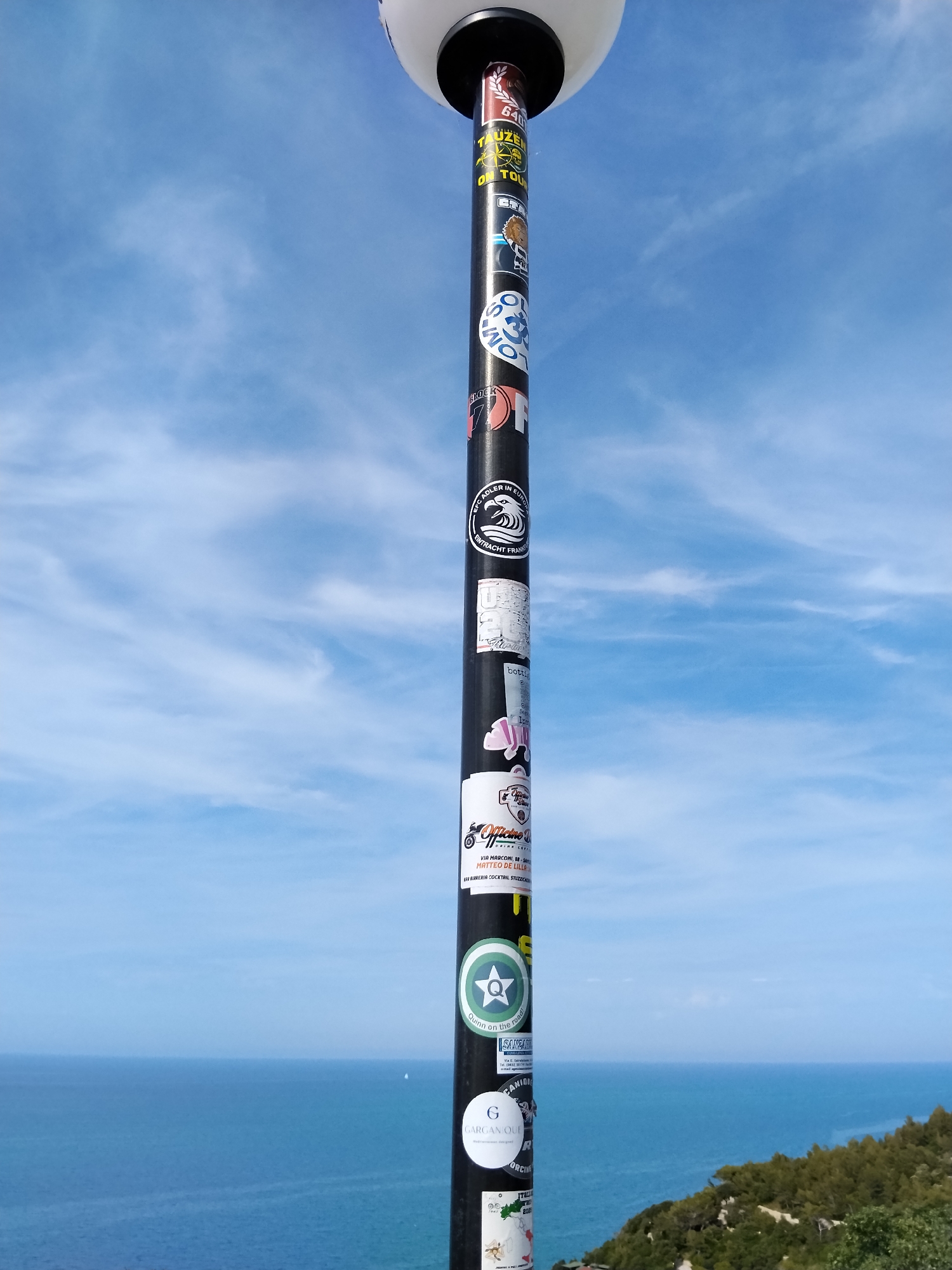 A tall, black metallic pole, topped with a spherical white light fixture, occupies the right foreground of the image. The pole is extensively covered with various stickers of different shapes, sizes, and colors. Notable stickers from top to bottom include: a yellow sticker with "TAUZEN ON TOUR," a round blue sticker with "S-33," a black sticker featuring a white wave logo and "EFC ADLER IN EUROPA" with "ENTRACHT FRANKFURT," a white sticker with a motorcycle graphic and text "Officine 'A MATTEO DE LILLO' DRINK FOOD VIA MARCONI, 33 - VICO DEL GARGANO," a green circular sticker with a white 'Q' and star labeled "Quinn on the road!", and a white circular sticker with a black 'G' and "GARGANO LIFE Mediterranean designed."
The background consists of a clear blue sky with sparse, wispy white clouds. Below the sky, a vast expanse of calm, deep blue sea stretches horizontally, meeting a distant horizon. A small white sailboat is discernible on the sea towards the left. On the lower right, a green, treed hillside or cliff descends towards the sea. No people are visible in the scene. The presence of the "Vico del Gargano" text on one of the stickers confirms the location as Vico del Gargano, Italy.