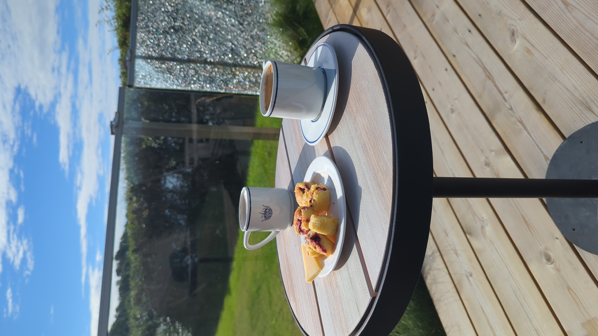 A round, wooden-topped table with a black metal base stands on a wooden deck. On the table are two white mugs with blue trim. One mug contains coffee. Next to the mugs is a white plate with several small, golden pastries topped with red fruit filling and a slice of pale yellow cheese. In the background, a glass railing overlooks a green lawn and trees under a blue sky with white clouds. The scene is located in Larkollen, Norway.