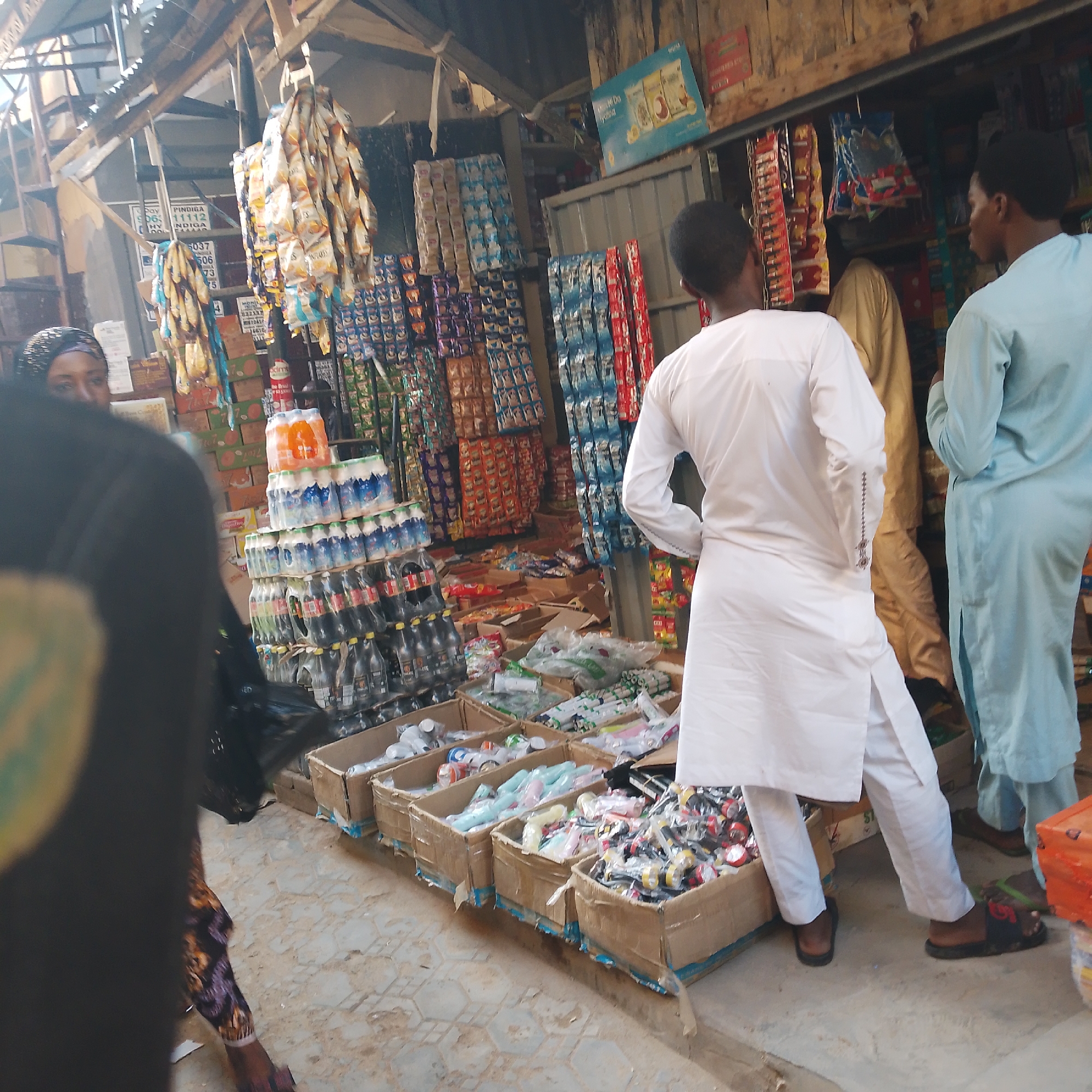 The image depicts an outdoor market scene in Arawa, Nigeria. In the foreground, the back of a person wearing a black abaya is visible on the left. To their right, a person in a white kaftan stands, facing away from the camera and towards a stall. Another person, partially visible, wears a light blue kaftan. Behind them, a man in a tan kaftan is also present. The market stall is stocked with a variety of goods, including bottles of beverages, packaged snacks, and other assorted items displayed in boxes and on shelves. The overall environment appears to be an open-air market with a corrugated metal roof structure overhead. The ground is paved with concrete tiles. The scene conveys a typical market day in a Nigerian city.