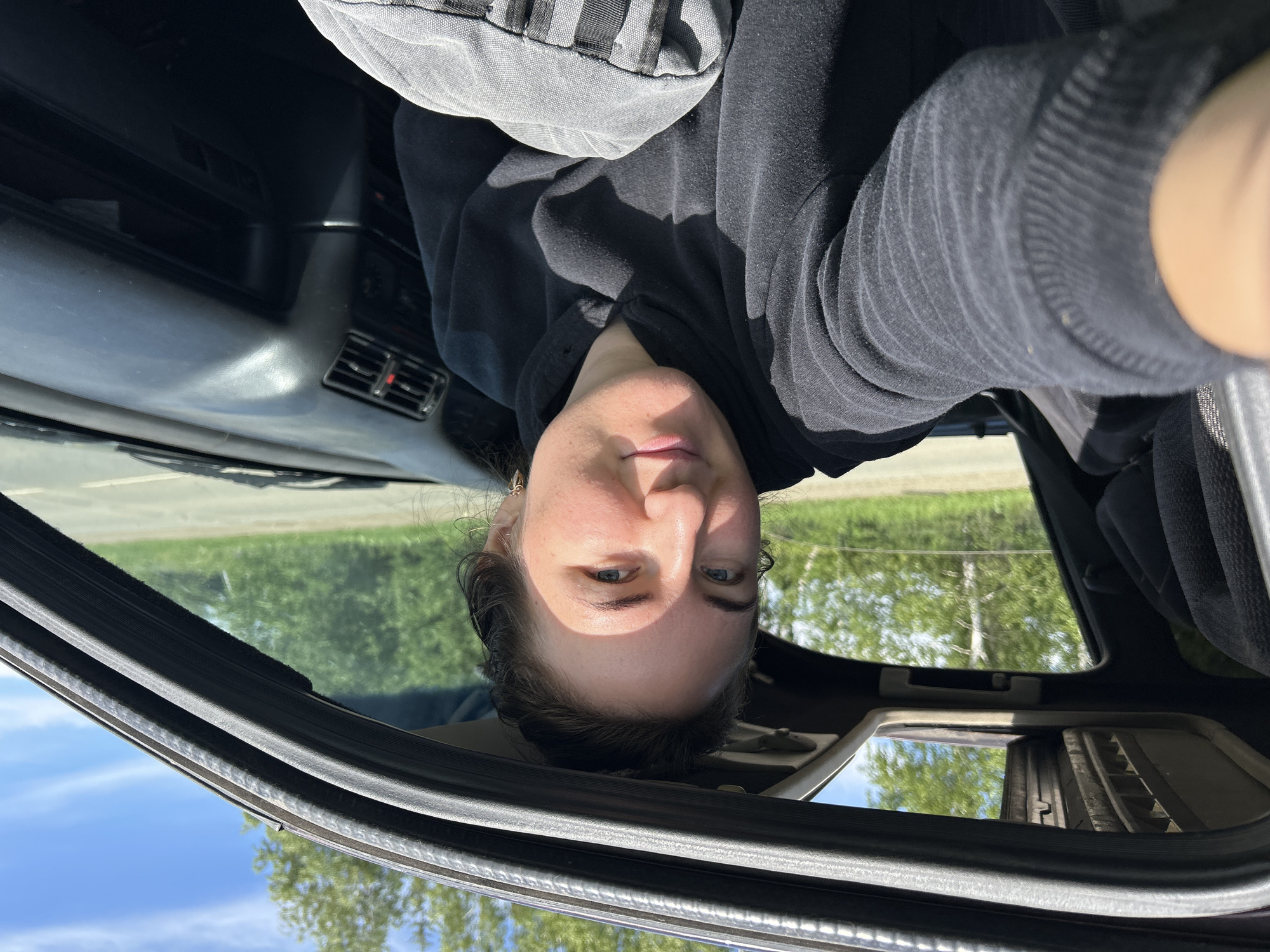 In Shabury, Russia, a woman with brown hair pulled back is captured in a selfie taken from inside a car. She is wearing a black hoodie and looking towards the camera with a gentle smile. The car's sunroof is open, revealing a bright blue sky with scattered clouds and green trees in the background. Sunlight illuminates her face, highlighting her features. The interior of the car is visible, with the dashboard and part of the passenger seat on the right side. The woman appears to be in a relaxed and comfortable setting, possibly on a journey or taking a break during a drive.