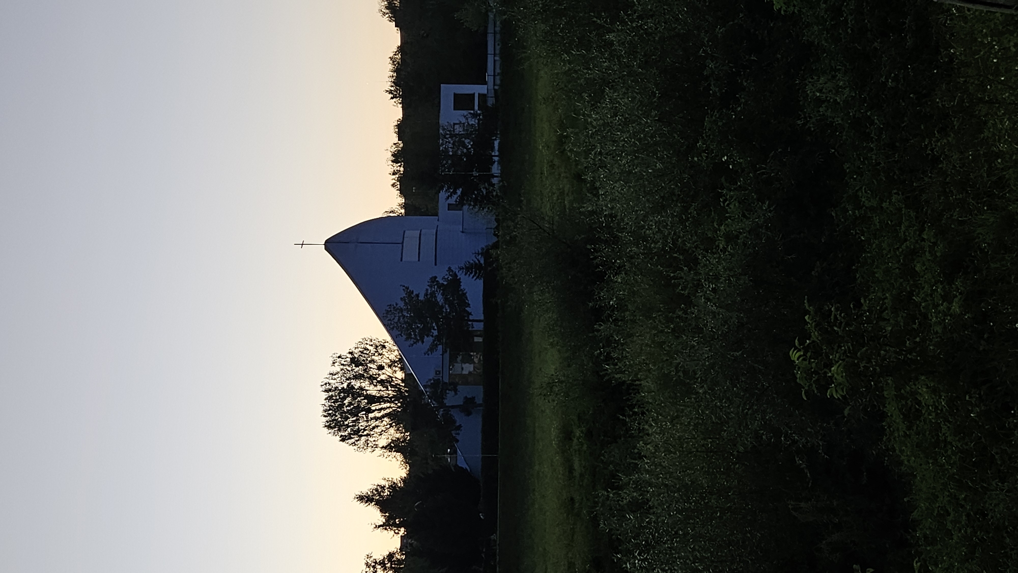 The image shows a modern, light blue church building with a distinctive triangular roof and a cross on top, silhouetted against a pale sky during what appears to be dusk. The church is situated behind a line of trees, and in the foreground, there is a dense area of dark green vegetation, possibly a field or a park. The architecture of the church suggests a contemporary design, possibly built in the late 20th or early 21st century. Without further context or distinguishing landmarks within the image itself, it is not possible to definitively identify the specific location within the city of Prešov, Slovakia. The scene is quiet and serene, with no people or visible activity. The lighting indicates either early morning or late evening.