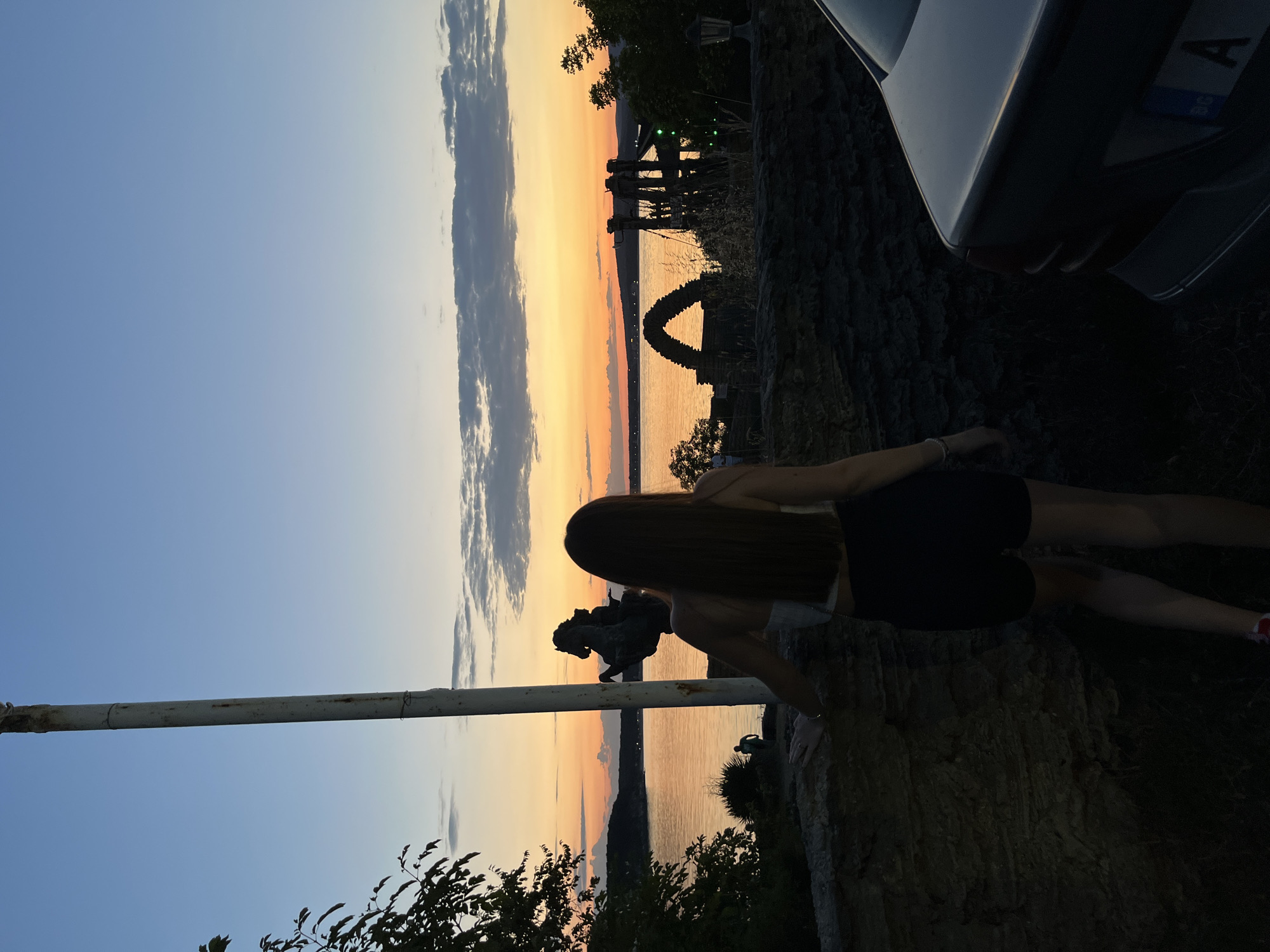 A young woman with long, brown hair stands with her back to the camera, facing a sunset over a body of water. She is wearing a white top and black shorts. In the foreground, a low stone wall is visible, with a portion of a car on the right side of the frame. Behind the woman and to her left, a tall pole stands prominently. The water reflects the warm colors of the sunset, with a silhouette of trees and structures on the opposite shore. The sky transitions from a soft blue at the top to shades of orange and yellow near the horizon. The scene appears to be a scenic overlook, possibly in Kiten, Bulgaria, given the presence of a nearby arch structure reminiscent of historical ruins often found in coastal Bulgarian towns.