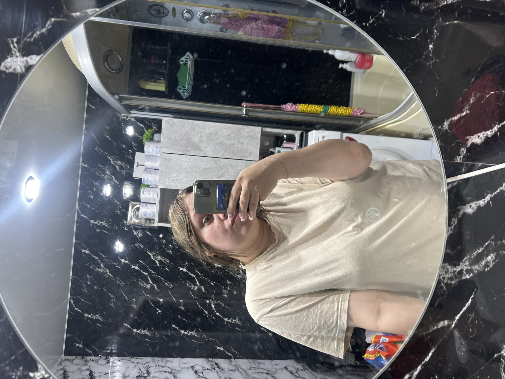 A woman is taking a selfie in a bathroom mirror. The bathroom has black and white marble-patterned walls. A white washing machine is visible behind her, as well as a cabinet with various items. The location is the city of Ольховка, Russia.