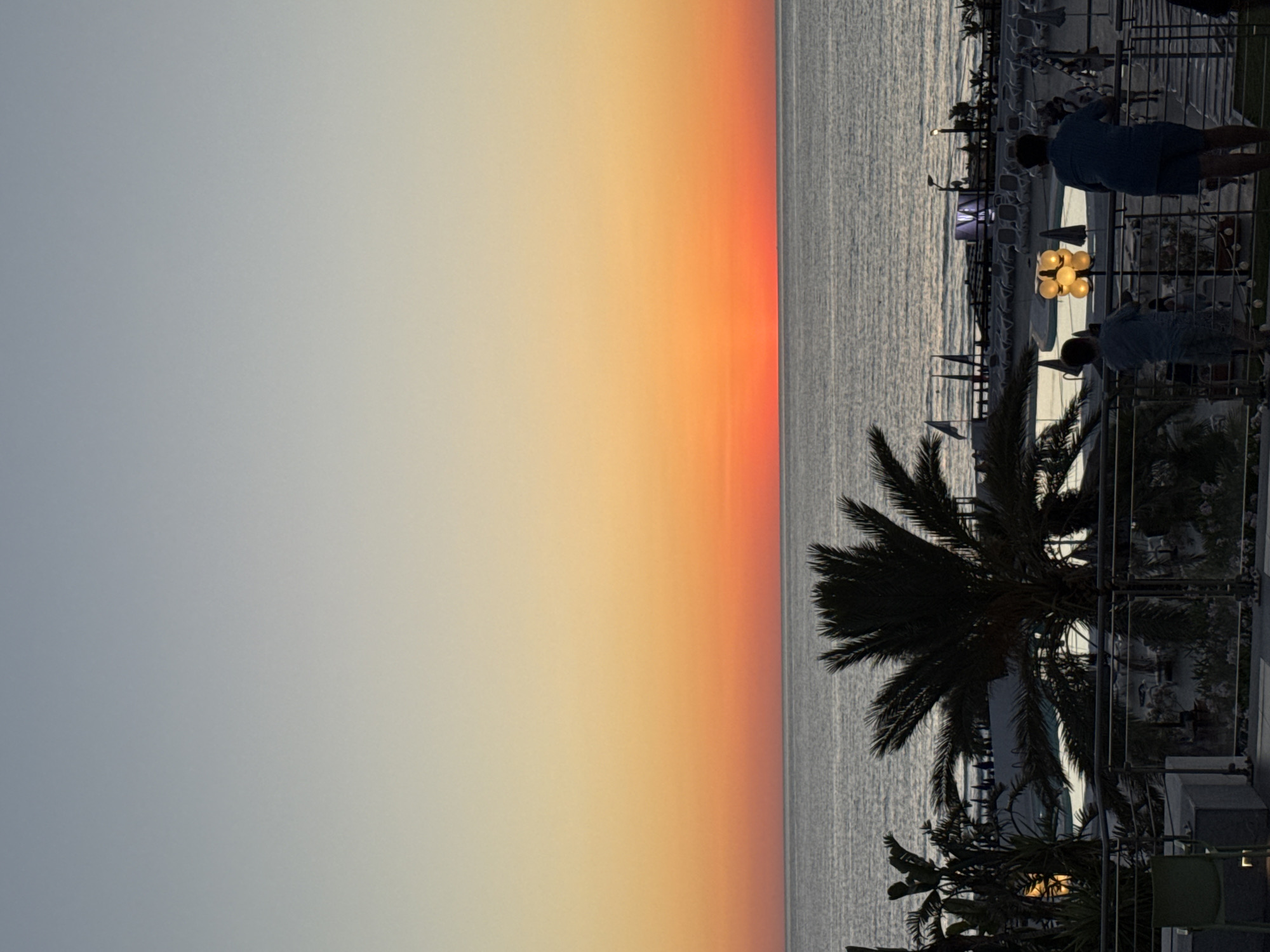 The image captures a serene sunset over the Mediterranean Sea, viewed from a vantage point in Isola delle Femmine, Italy. The sky is a gradient of soft blues at the top, transitioning through warm oranges and yellows near the horizon. The sea is calm, reflecting the sky's colors. In the foreground, silhouetted palm trees frame the view. Below, a resort area is visible with lounge chairs, a swimming pool, and lighting. Two individuals, one adult and one child, stand on a balcony, looking out at the sea. The scene conveys a peaceful evening atmosphere.