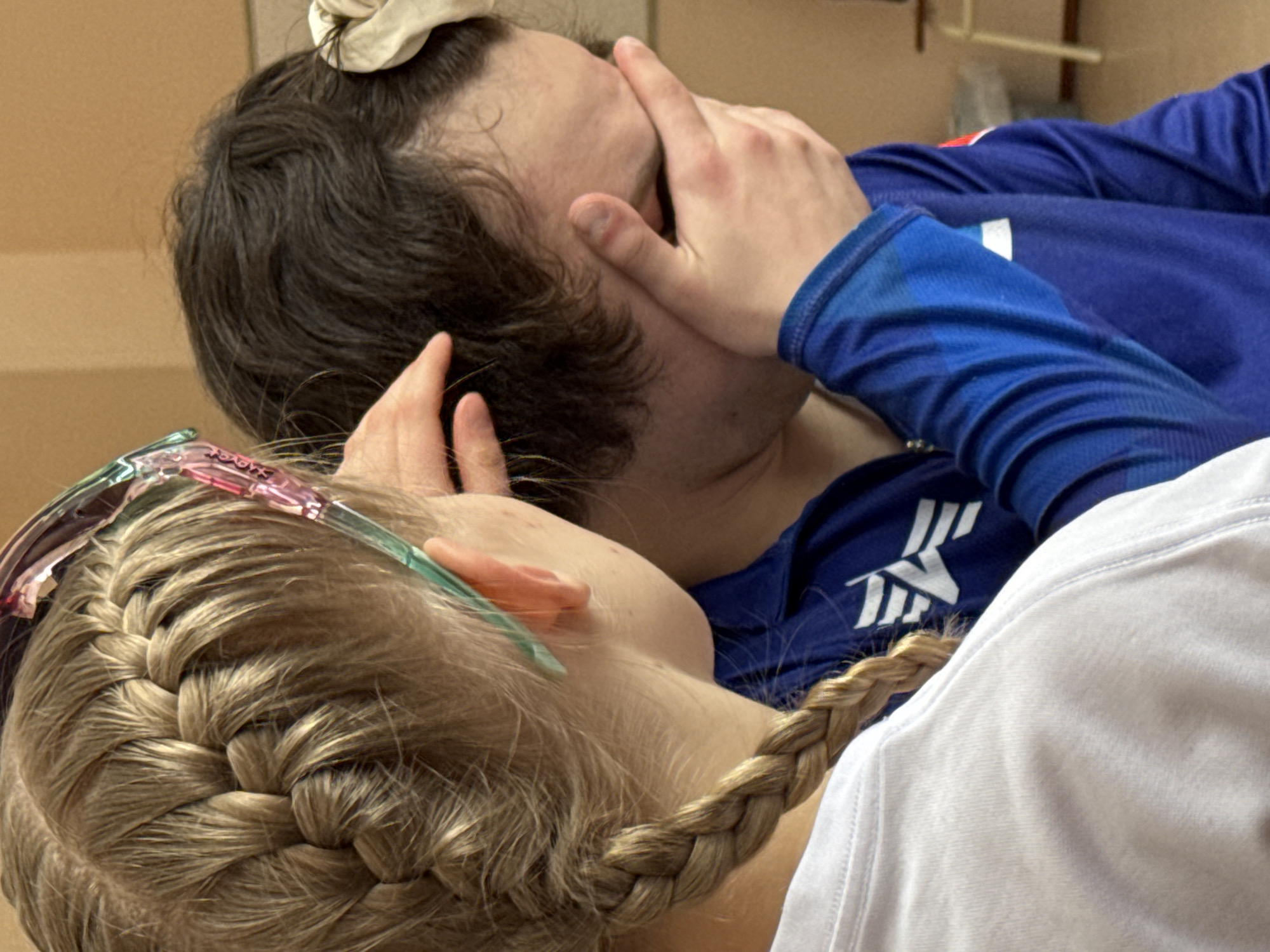 A photograph shows a close-up of two people lying down. A fair-skinned person with blond hair styled in a braid and pink and green hair accessories is close to the camera, with their head resting near the shoulder of a darker-haired person wearing a blue long-sleeved shirt with a white logo. The person in the blue shirt has their hands covering part of their face. The setting appears to be indoors, given the wall color. There is no visible evidence of the location being in Berezniki, Russia.