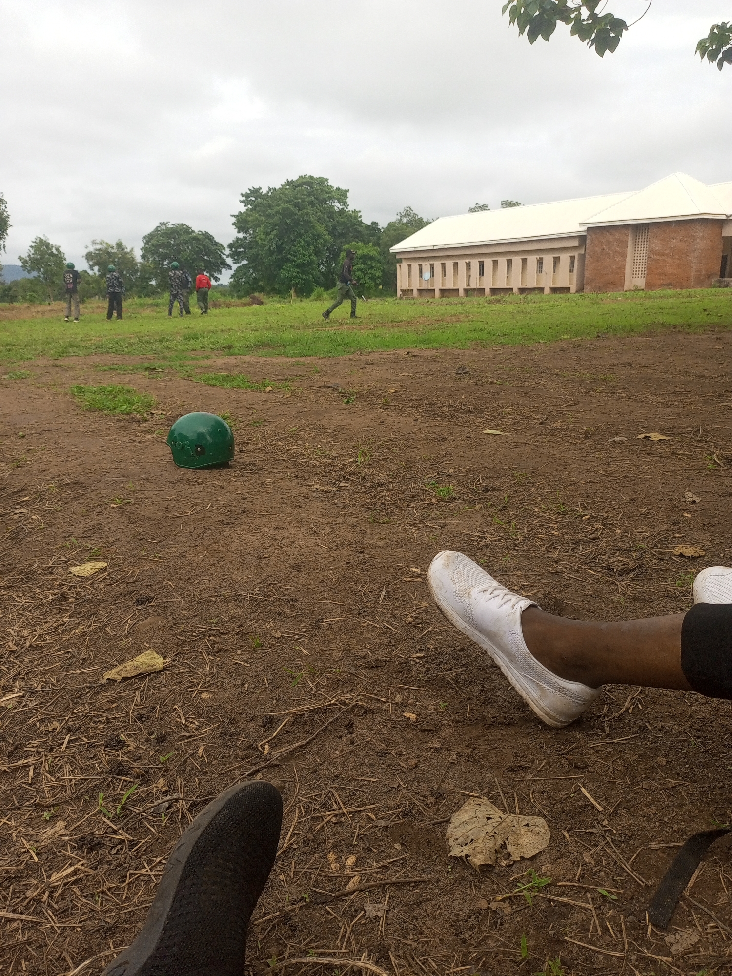 In Takum, Nigeria, on a cloudy day, a green helmet rests on dry, disturbed earth in the foreground, suggesting a training exercise or a recreational activity. In the middle ground, several individuals, possibly military personnel or cadets, are scattered across a grassy field. Some are wearing camouflage uniforms and helmets, while others are in different attire. One person in a red shirt stands out. In the background, a low-rise building with a white roof and cream-colored walls is visible, with a brick section on the right. The environment appears to be an open area with trees and sparse vegetation, hinting at a rural or semi-urban setting within Takum. The composition is grounded by a close-up of a person's legs and feet wearing white sneakers, suggesting the photographer is part of the scene or observing it from a close vantage point.