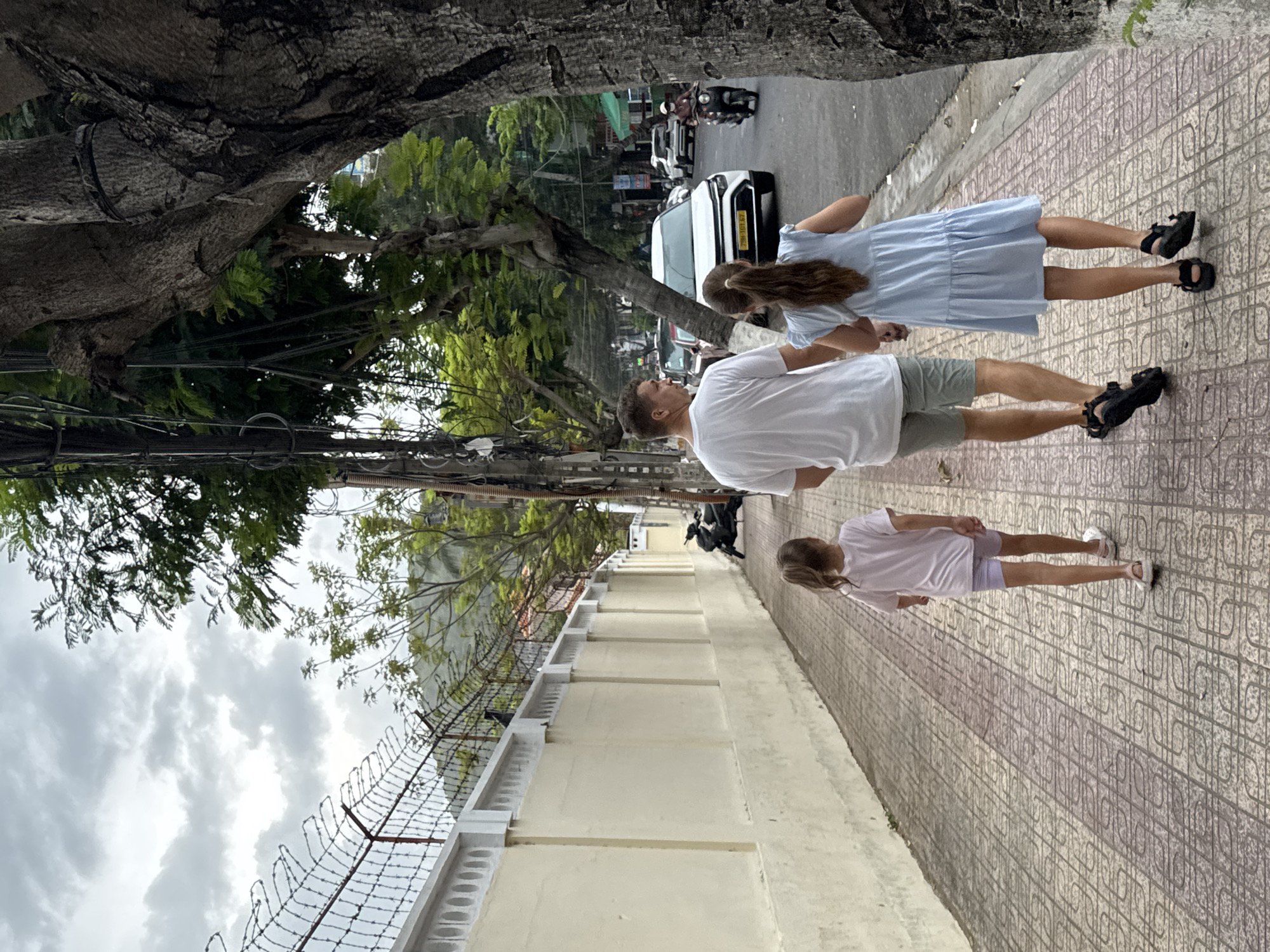 The photo captures a scene on a city sidewalk in Bac Nha Trang Ward, Vietnam. A man in a white shirt and khaki shorts walks alongside a girl in a blue dress and another girl in a white t-shirt and purple shorts. They are walking on a tiled sidewalk. To their left is a wall topped with a wire fence. Overhead are power lines and the branches of a large tree. The street is visible in the background with various vehicles. The sky is overcast.