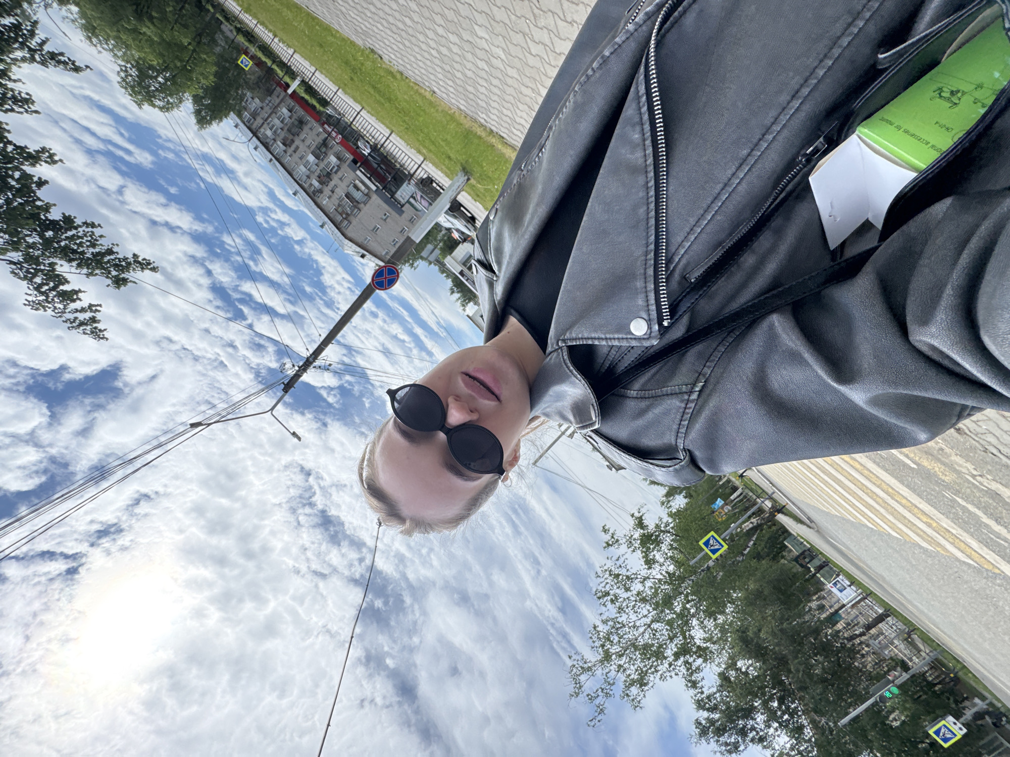 A woman with blond hair and dark sunglasses stands on a street in the city of Chaikovsky, Russia, taking a selfie. She wears a black leather jacket with a green box and white paper sticking out of a pocket. Behind her, a sidewalk leads to a crosswalk with yellow pedestrian crossing signs. Trees line the street. A building stands across the street under a sky with fluffy white clouds. Telephone poles and wires are visible. A red and blue "no parking" sign is attached to a pole.