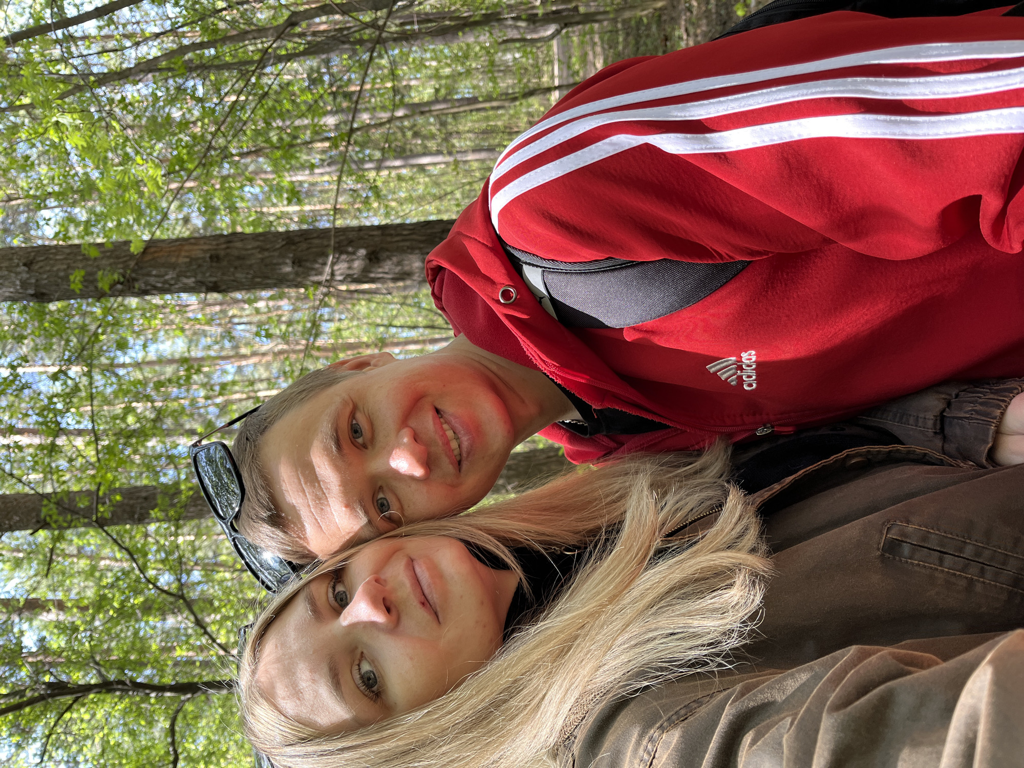 The image shows two people in a forest. A woman with blonde hair is positioned at the bottom of the frame wearing a brown jacket. Above her is a man wearing a red Adidas sweatshirt with white stripes. They are close to each other, and the background consists of trees and foliage. The location is Chaikovsky, Russia.